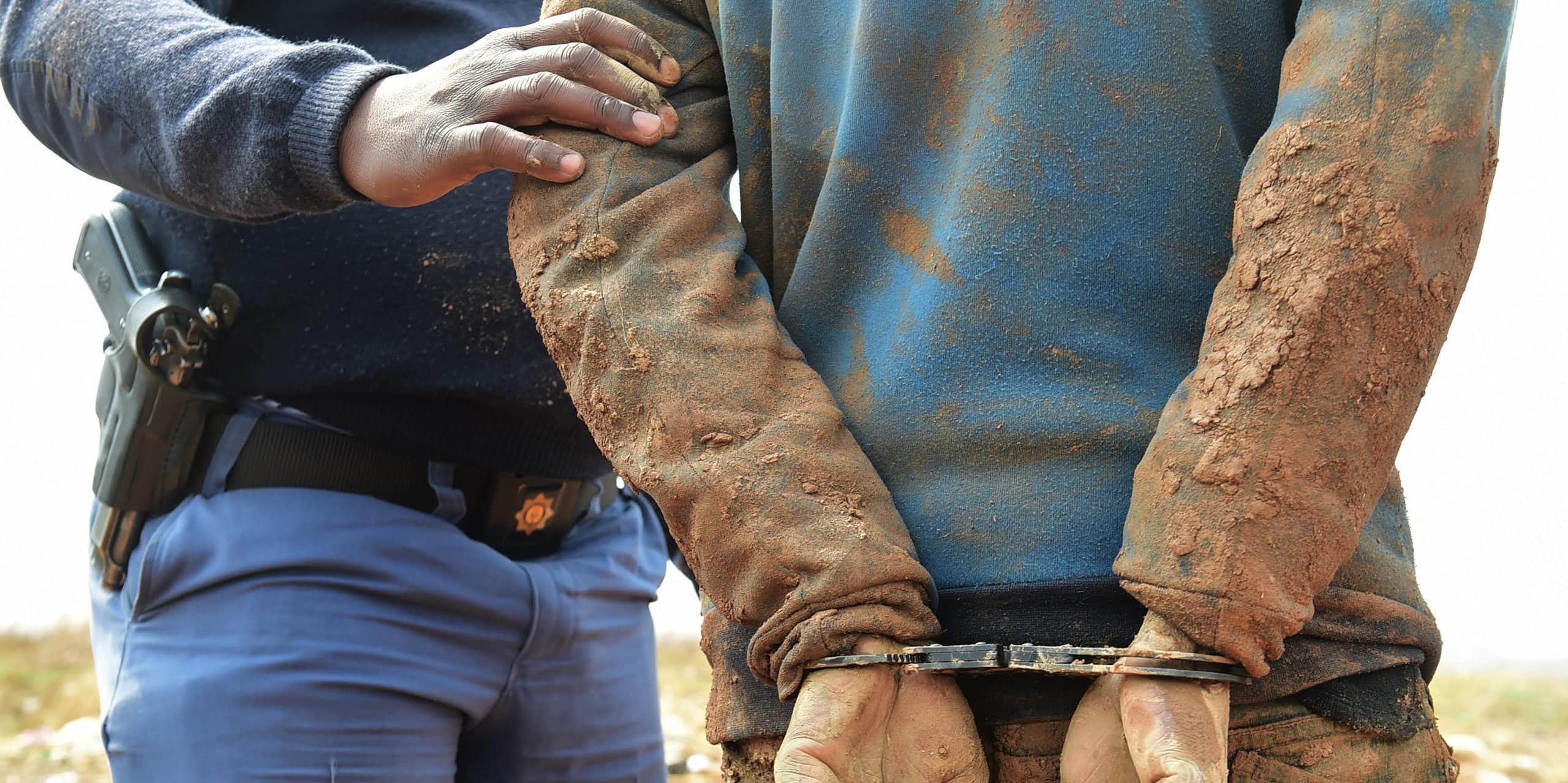 Seen from behind, a man in mud-caked clothing and his hands in handcuffs, while a man in blue uniform with a gun in a holster holds his arms