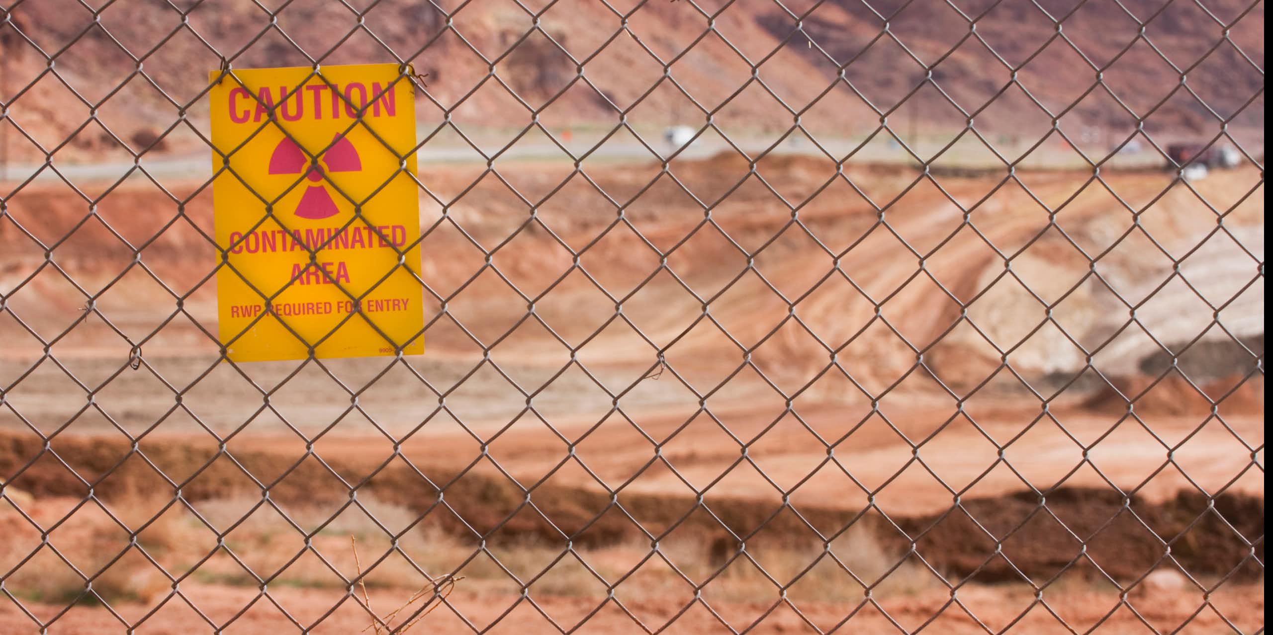 A big sandy area seen through a wire fence. Hanging on the fence is a notice - caution, contaminated area