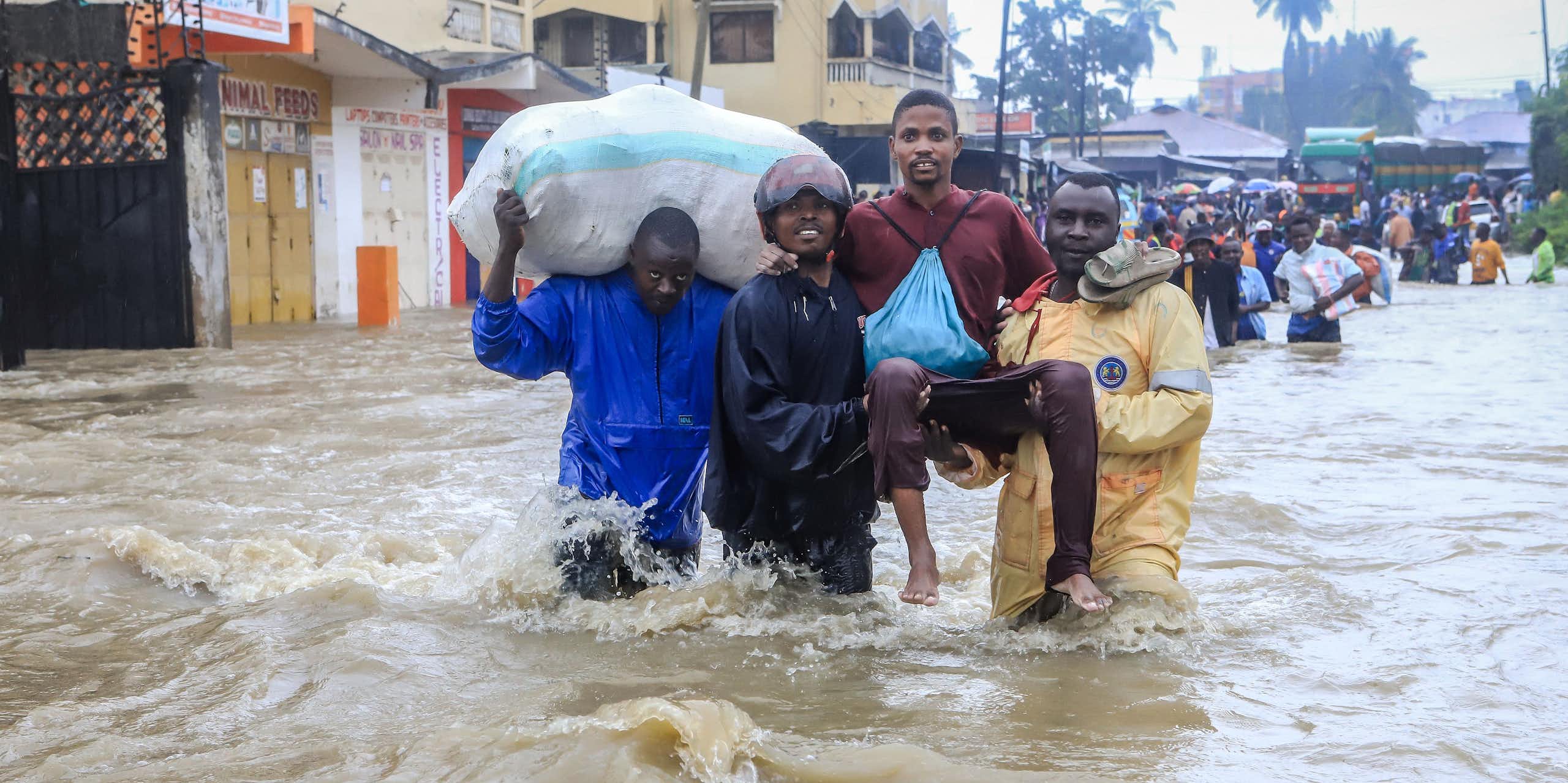 Two men carry another through a flooded city with water up to their waists while another man carries a giant sack. Behind them walk dozens of people, all wading through the water.