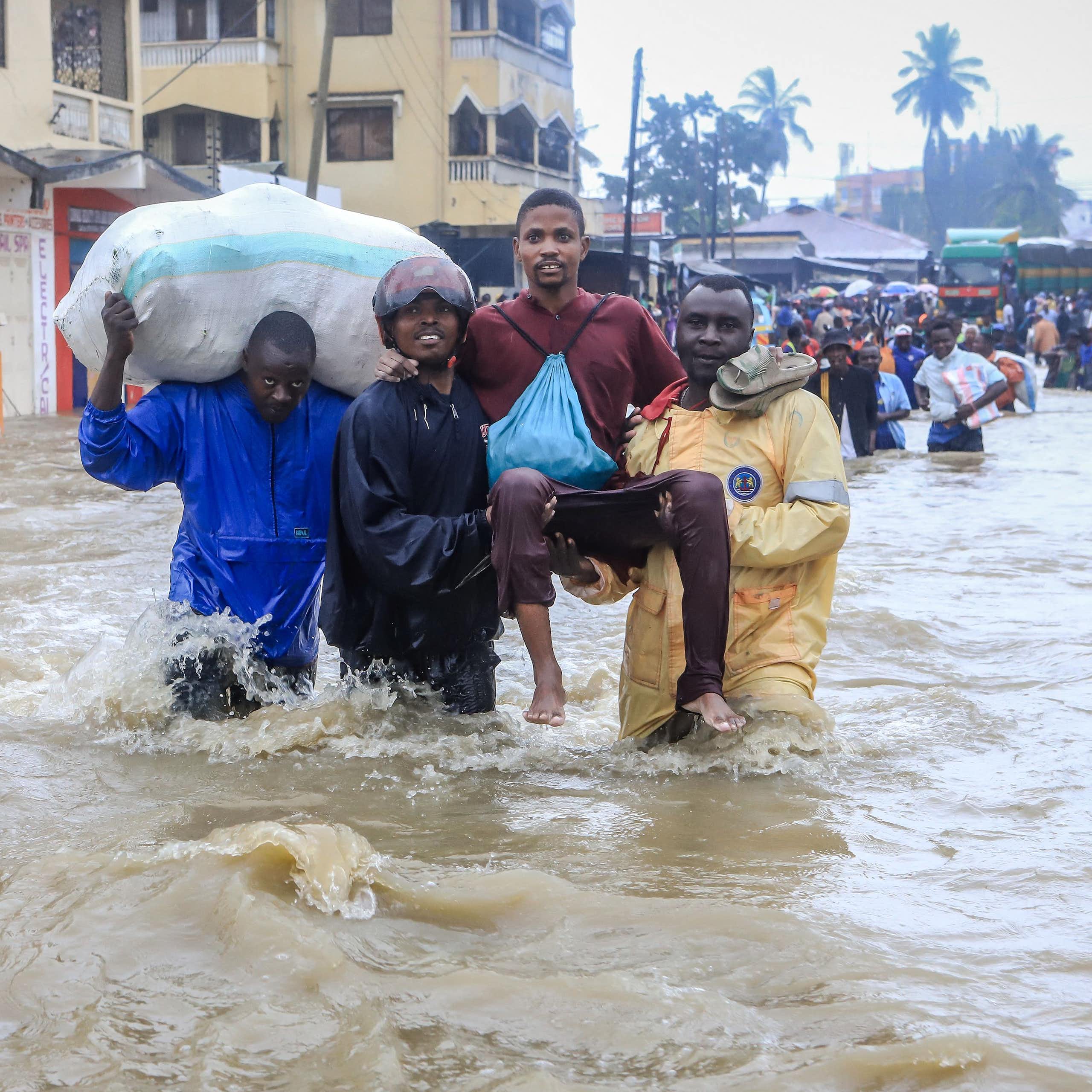 Two men carry another through a flooded city with water up to their waists while another man carries a giant sack. Behind them walk dozens of people, all wading through the water.