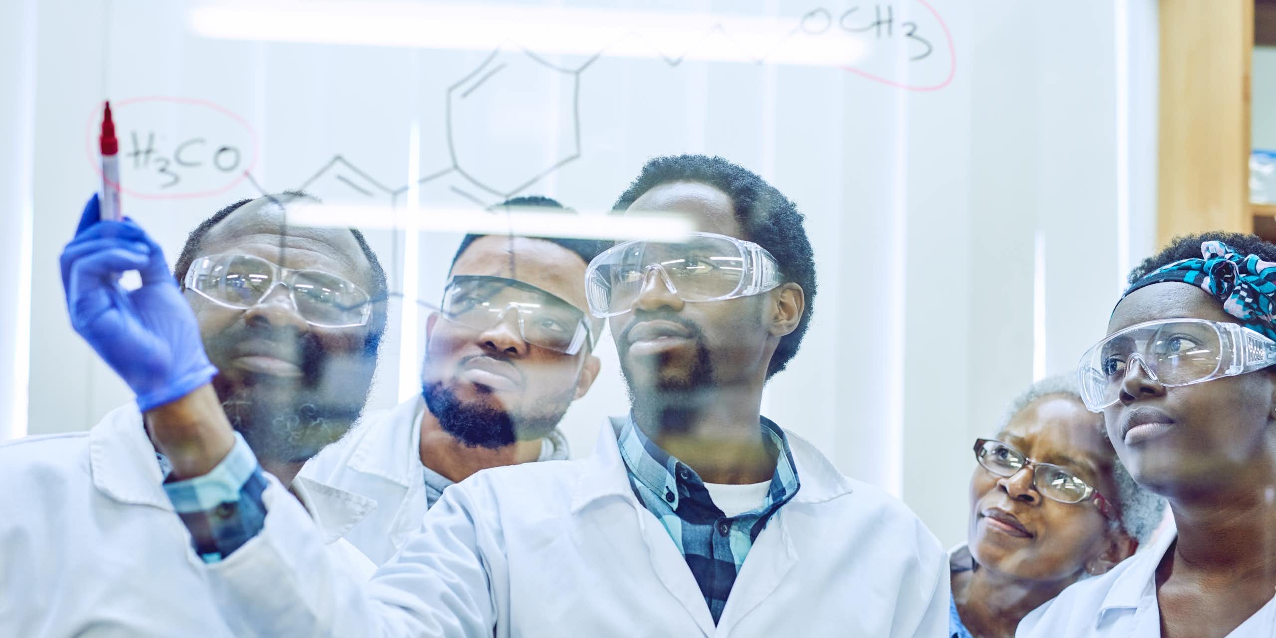 Five people in white coats and protective eyewear look at a clear wall on which one is writing a formula
