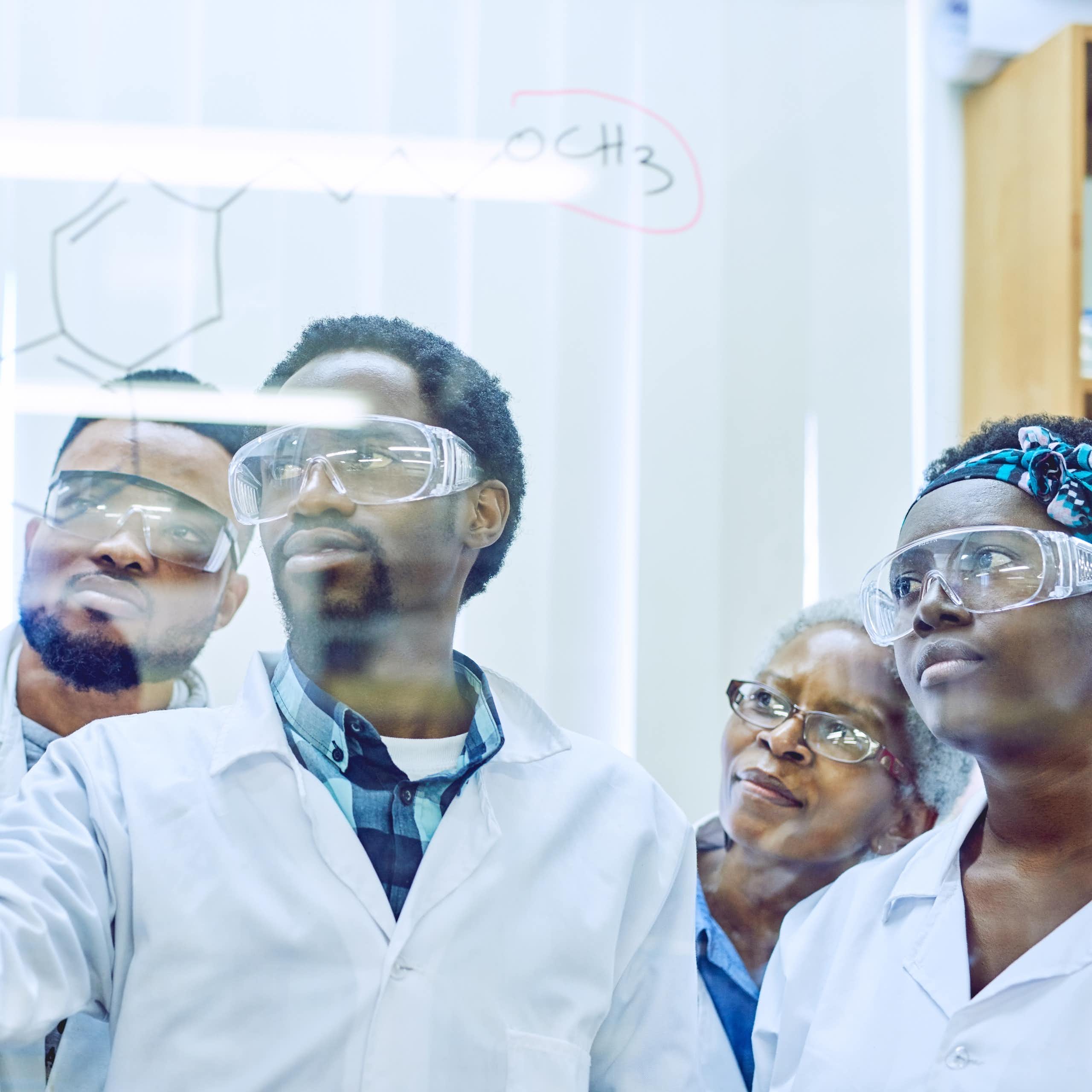 Five people in white coats and protective eyewear look at a clear wall on which one is writing a formula