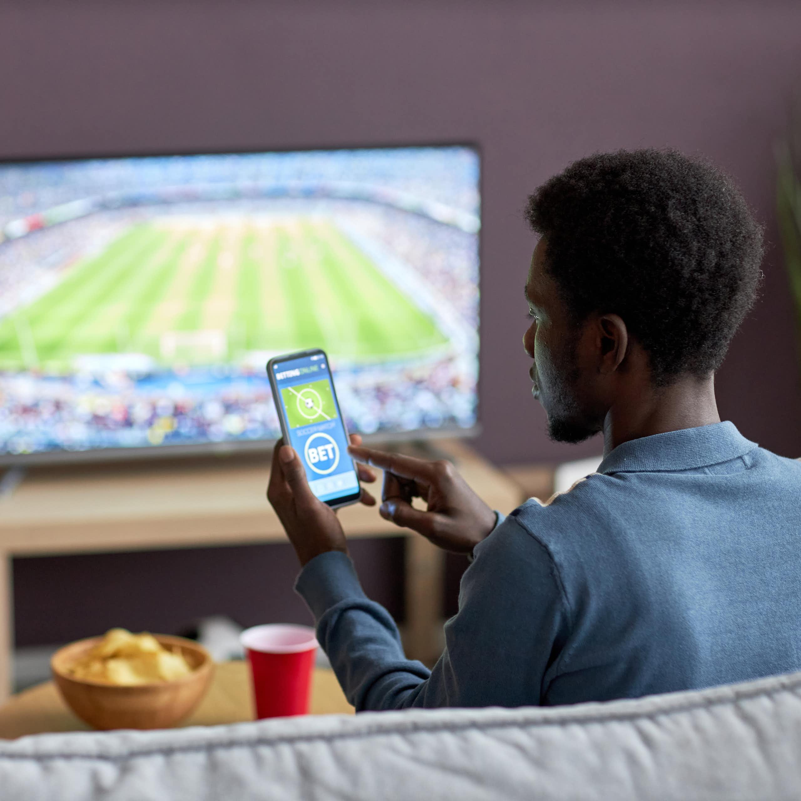 A man watching a sports event on television and placing a bet on his phone
