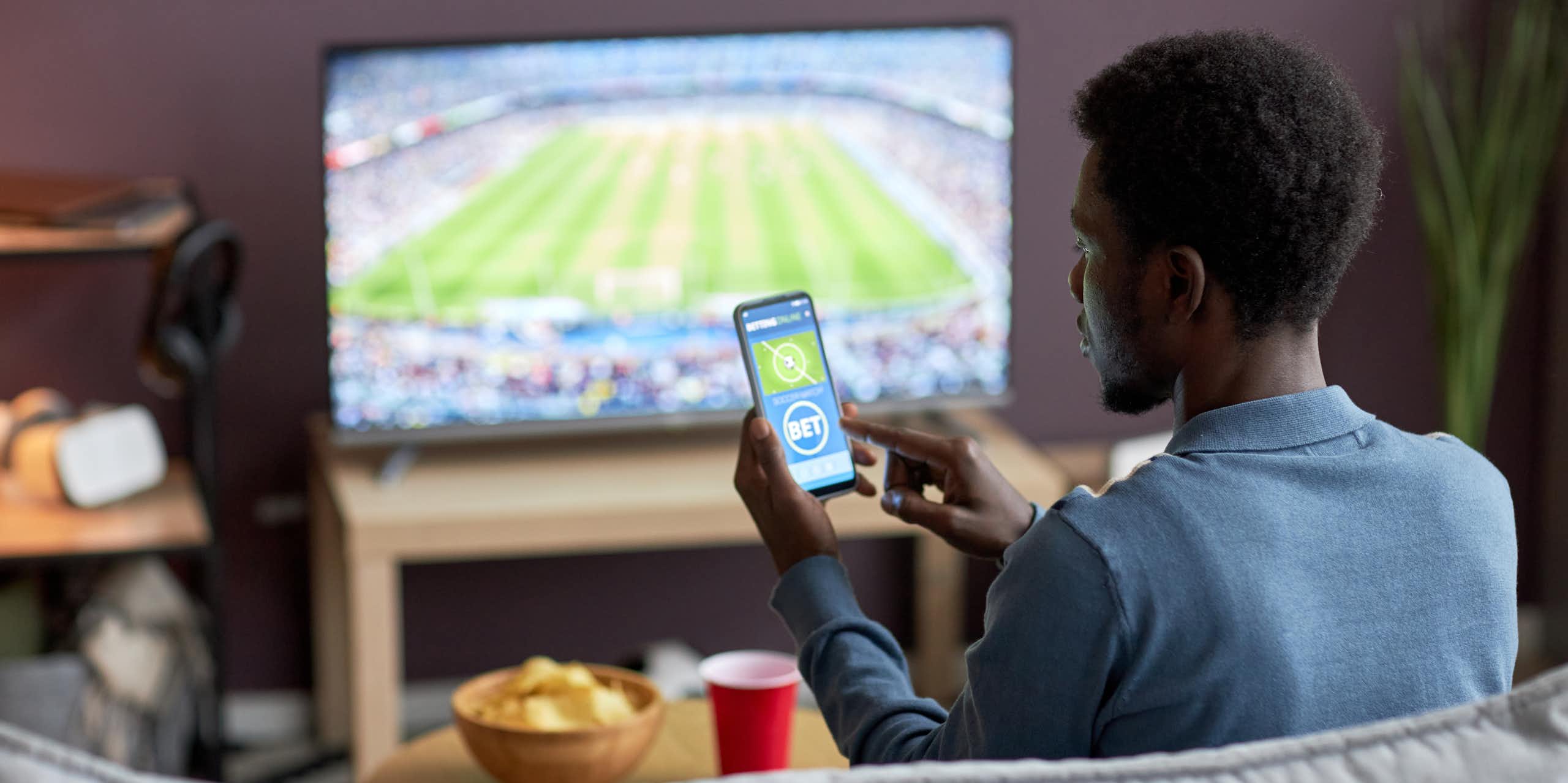 A man watching a sports event on television and placing a bet on his phone