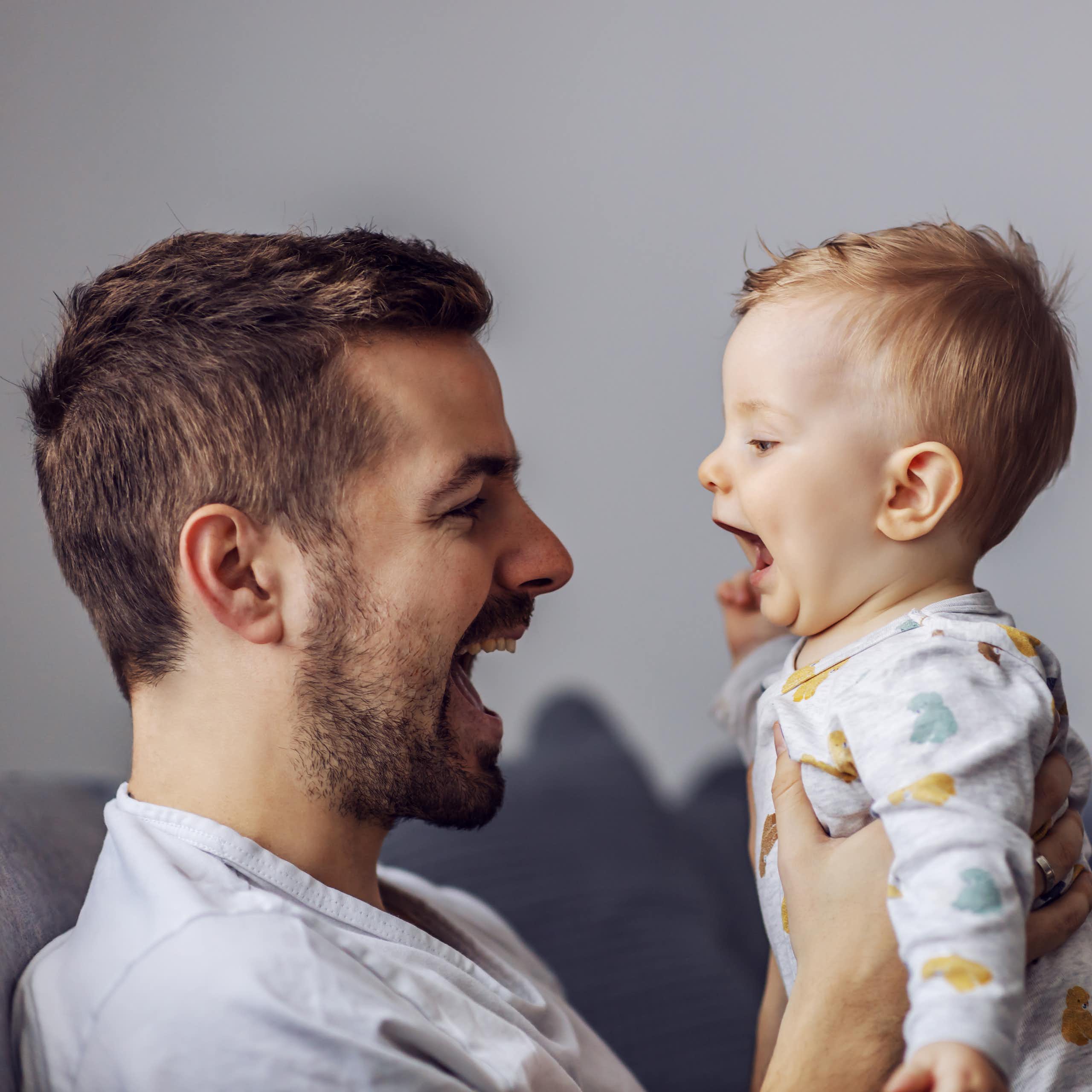 A man holding up a baby, both have their mouths open  talking to each other