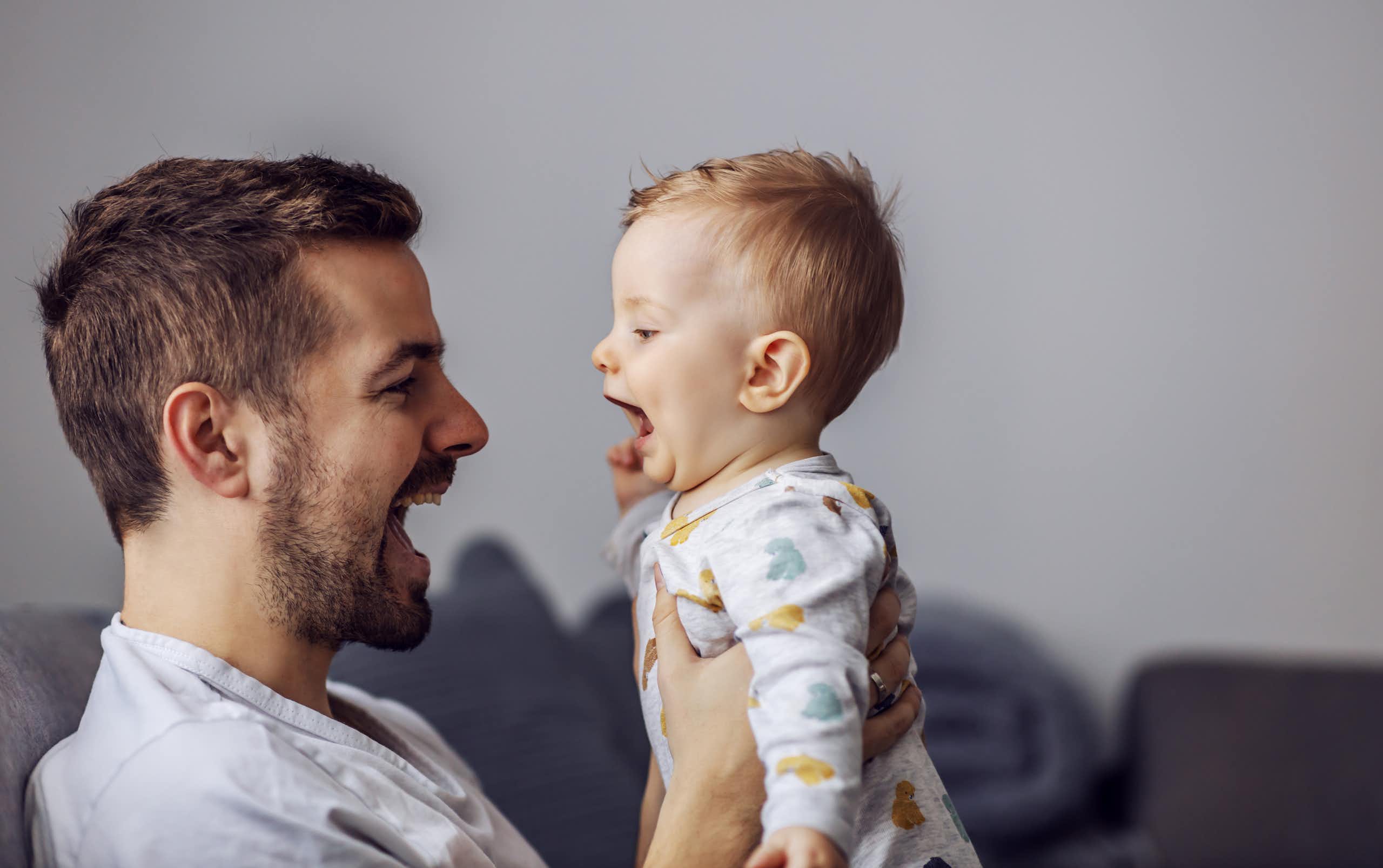 A man holding up a baby, both have their mouths open  talking to each other