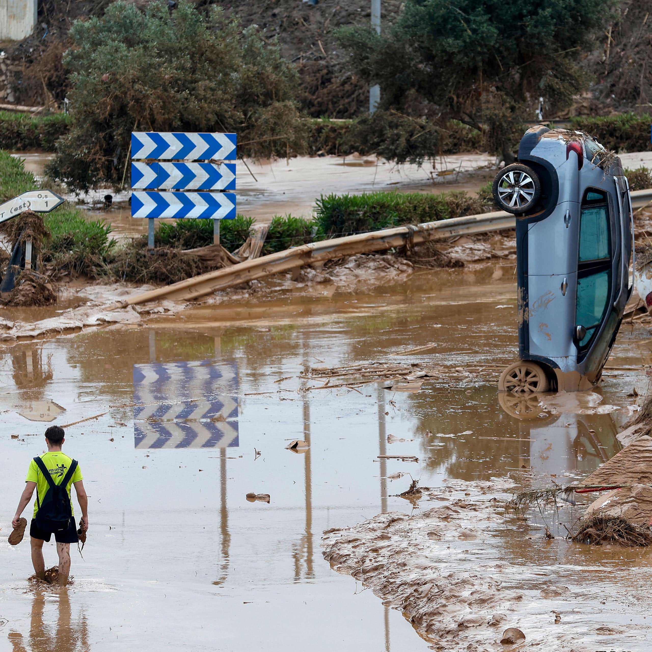 Duas pessoas caminham pela água lamacenta da enchente em uma estrada na Espanha, com carros destruídos ao fundo