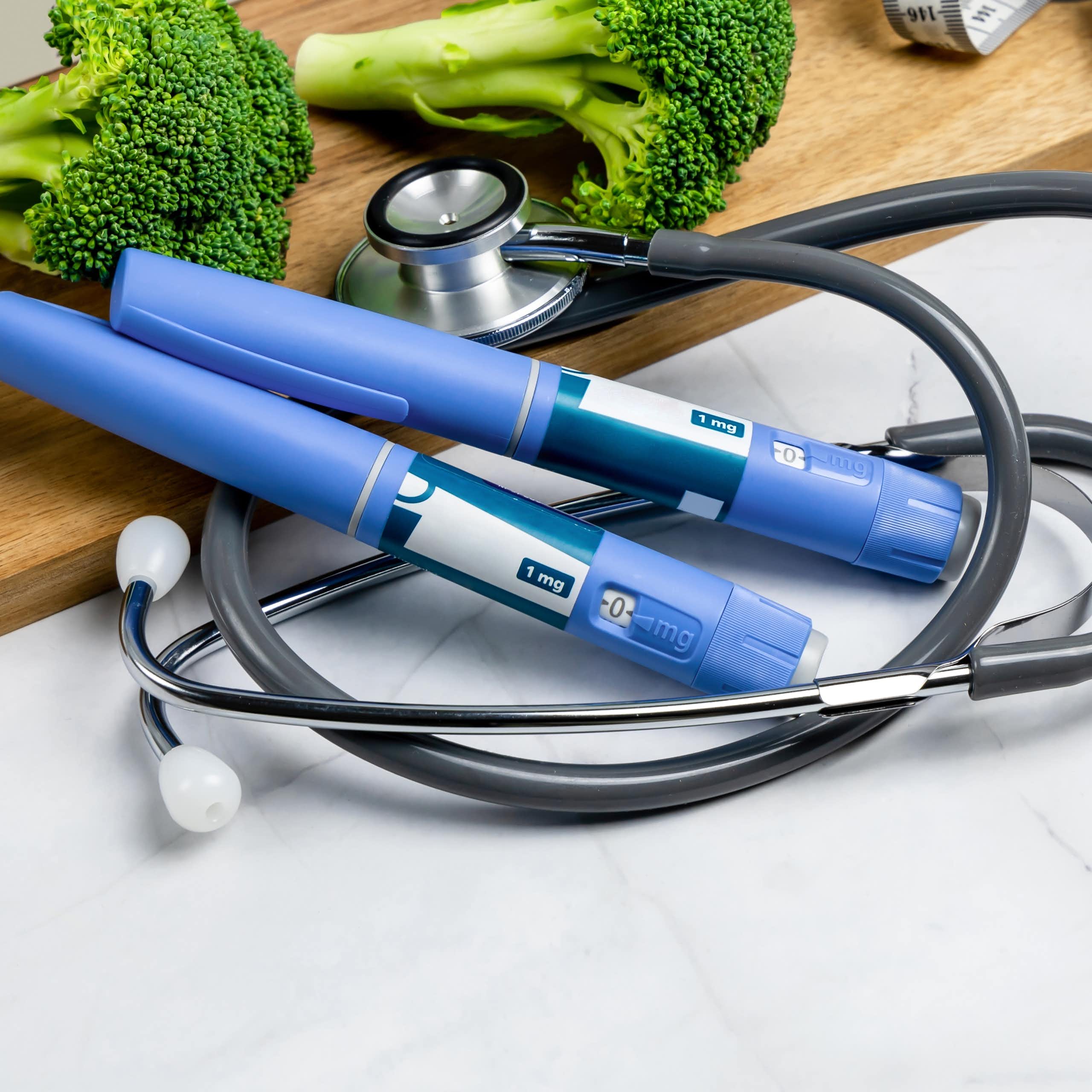 Two blue medication pens, a stethoscope, a measuring tape and some random broccoli
