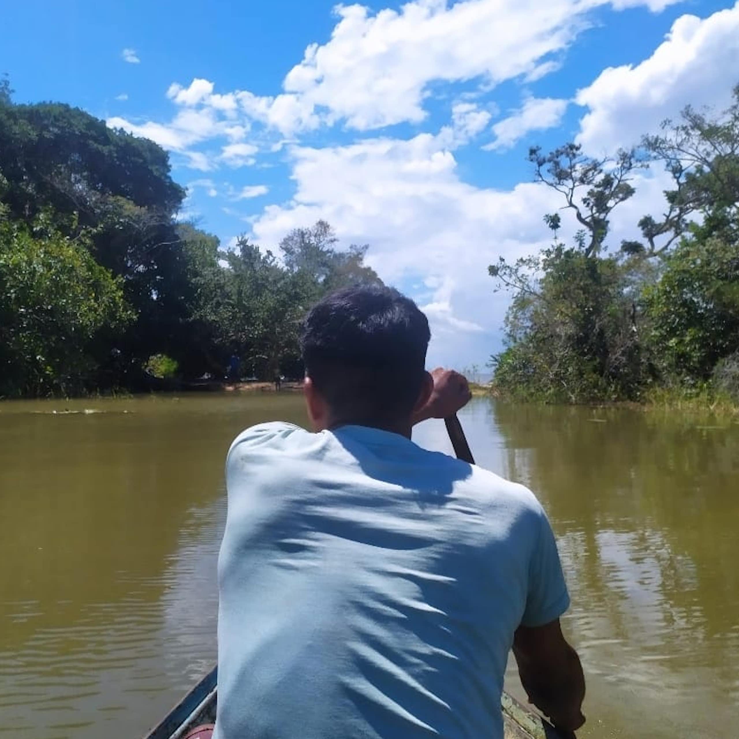 Foto mostra uma canoa sendo remada por um homem de costas. Ele está no centro do rio, e as margens dos dois lados são cobertas de florestas.