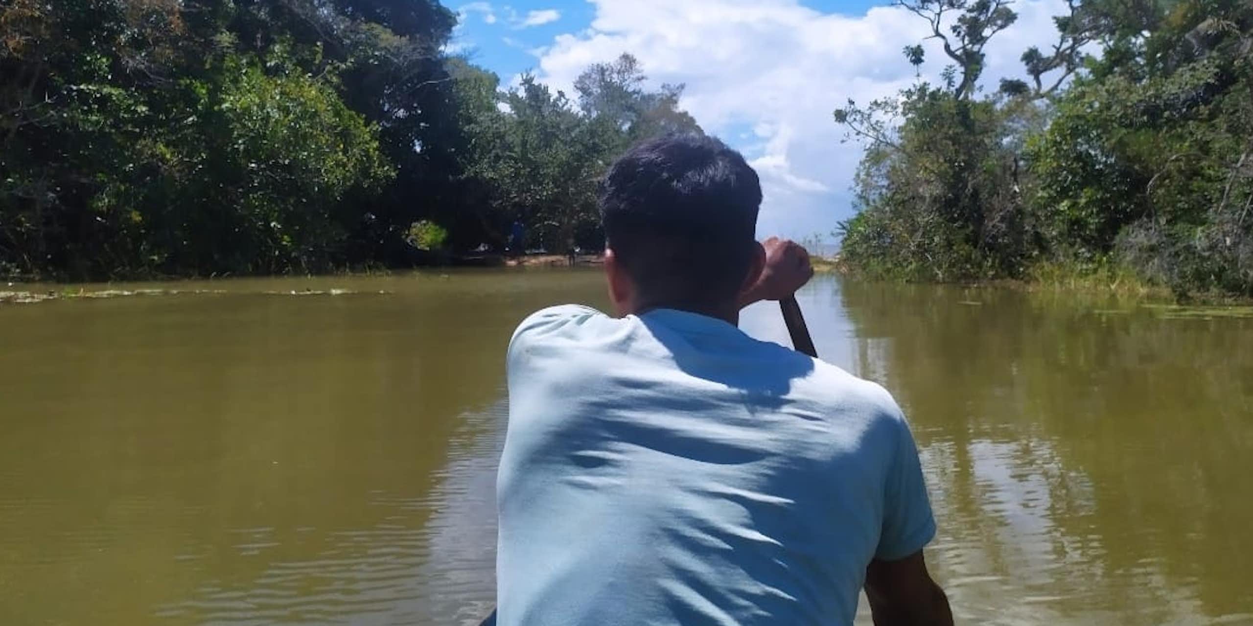 Foto mostra uma canoa sendo remada por um homem de costas. Ele está no centro do rio, e as margens dos dois lados são cobertas de florestas.