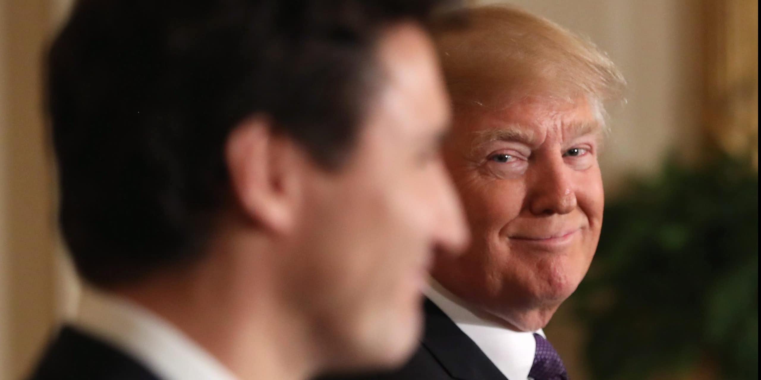 A man with orange-ish blond hair smiles over at a younger man with dark hair seen in silhouette.