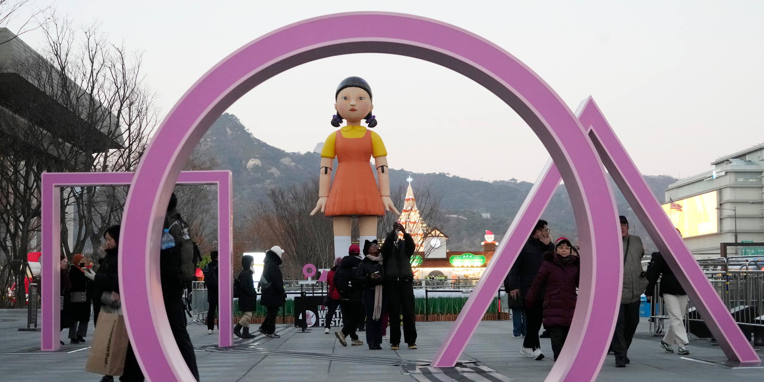 People walk past an art installation for Squid Games Season 2. It consists of a purple circle, square and triangle rising out of the ground in front of a giant doll with pigtails