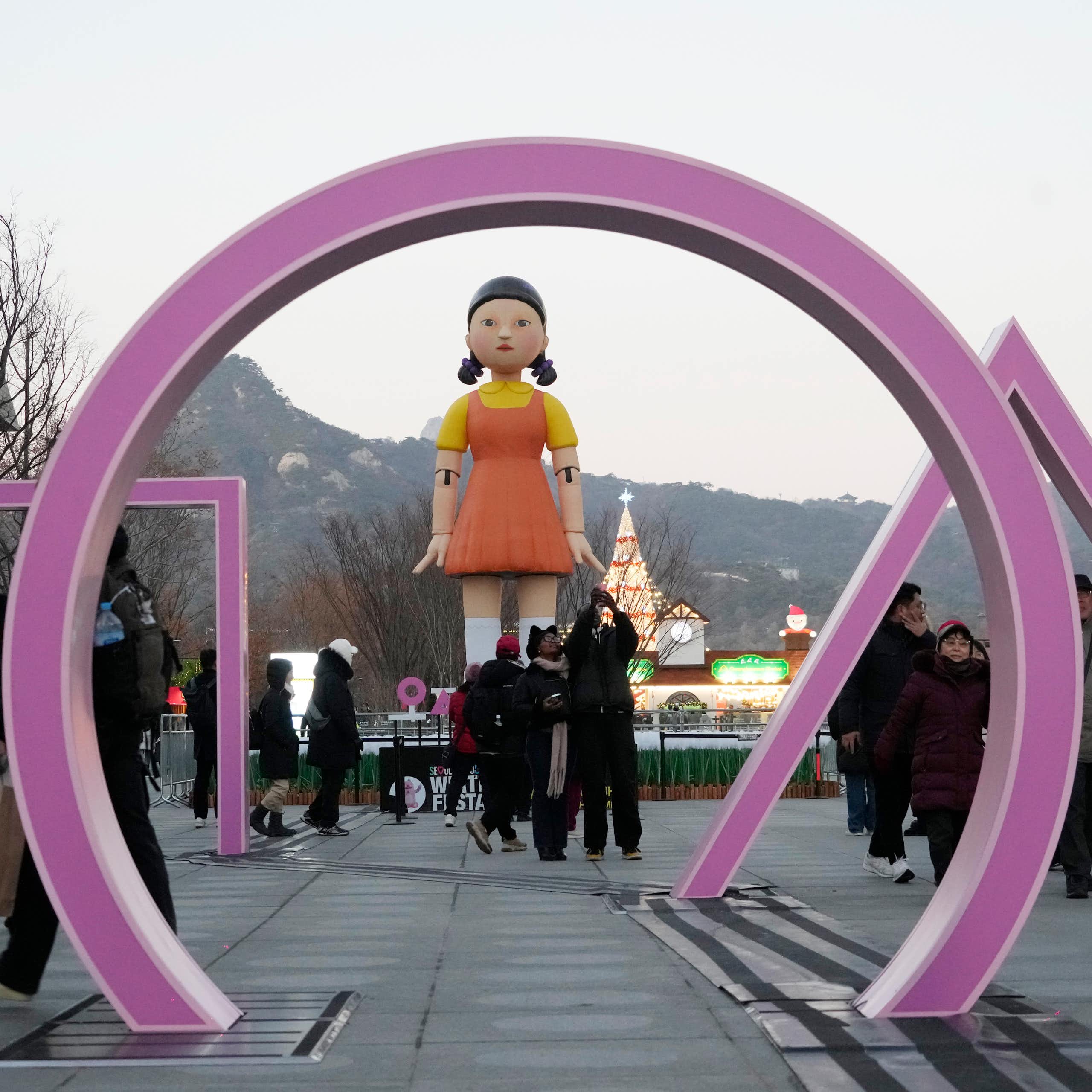 People walk past an art installation for Squid Games Season 2. It consists of a purple circle, square and triangle rising out of the ground in front of a giant doll with pigtails