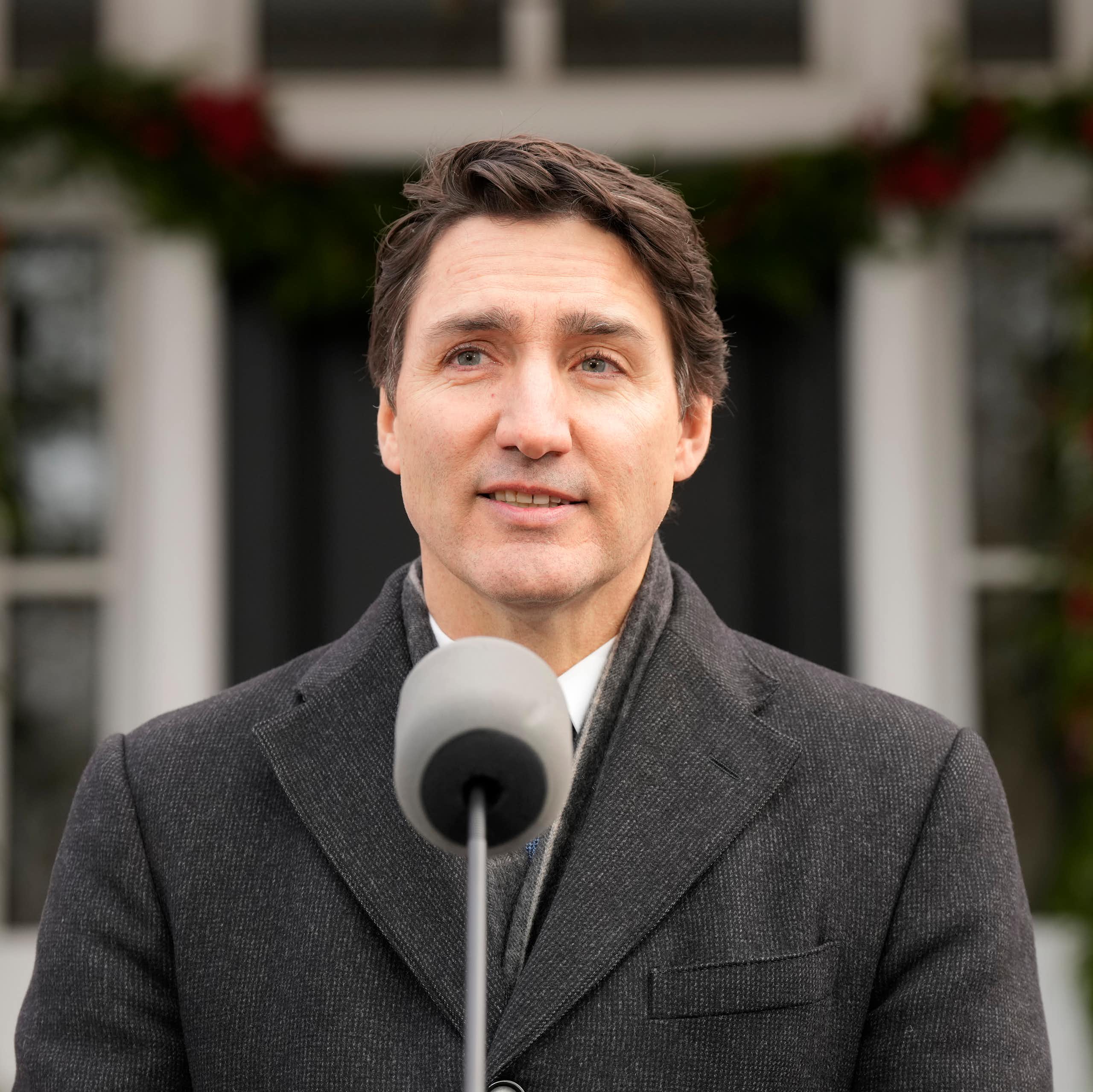 A dark-haired man in a grey winter coat speaks into a microphone with a faint smile on his face.