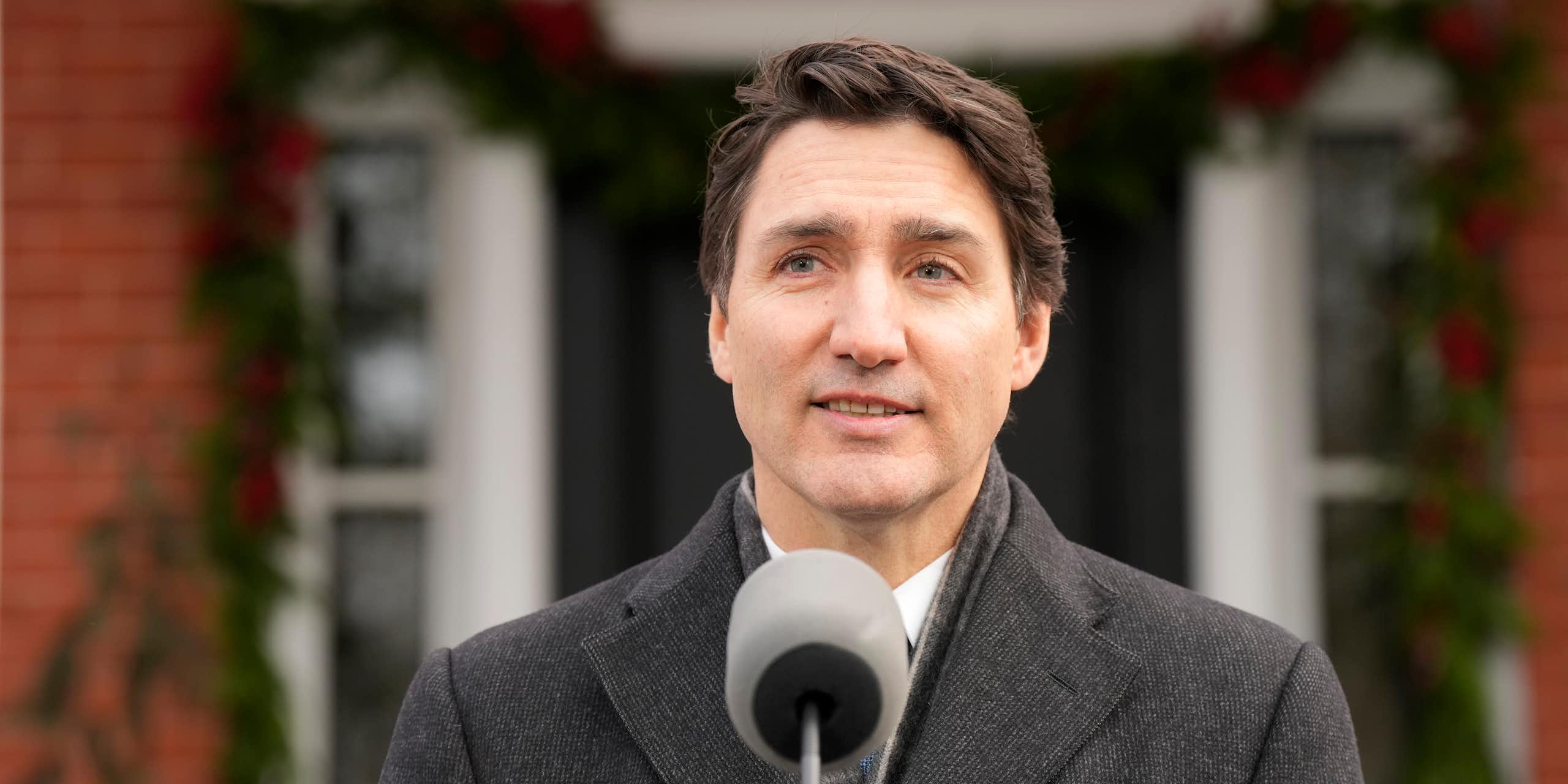 A dark-haired man in a grey winter coat speaks into a microphone with a faint smile on his face.