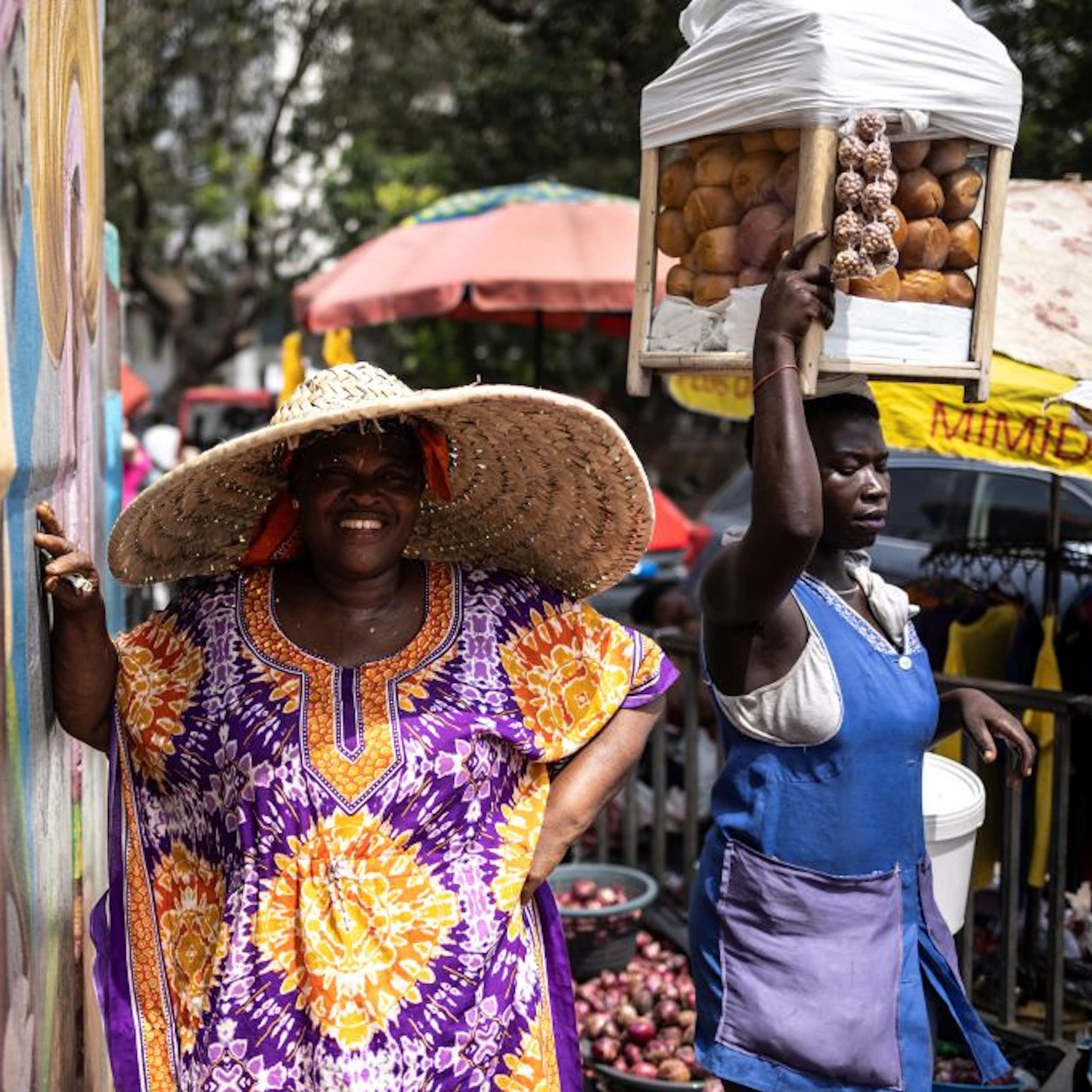 A smiling woman wearing a wide-brimmed hat. Another woman in the background with a container of goods on her head.