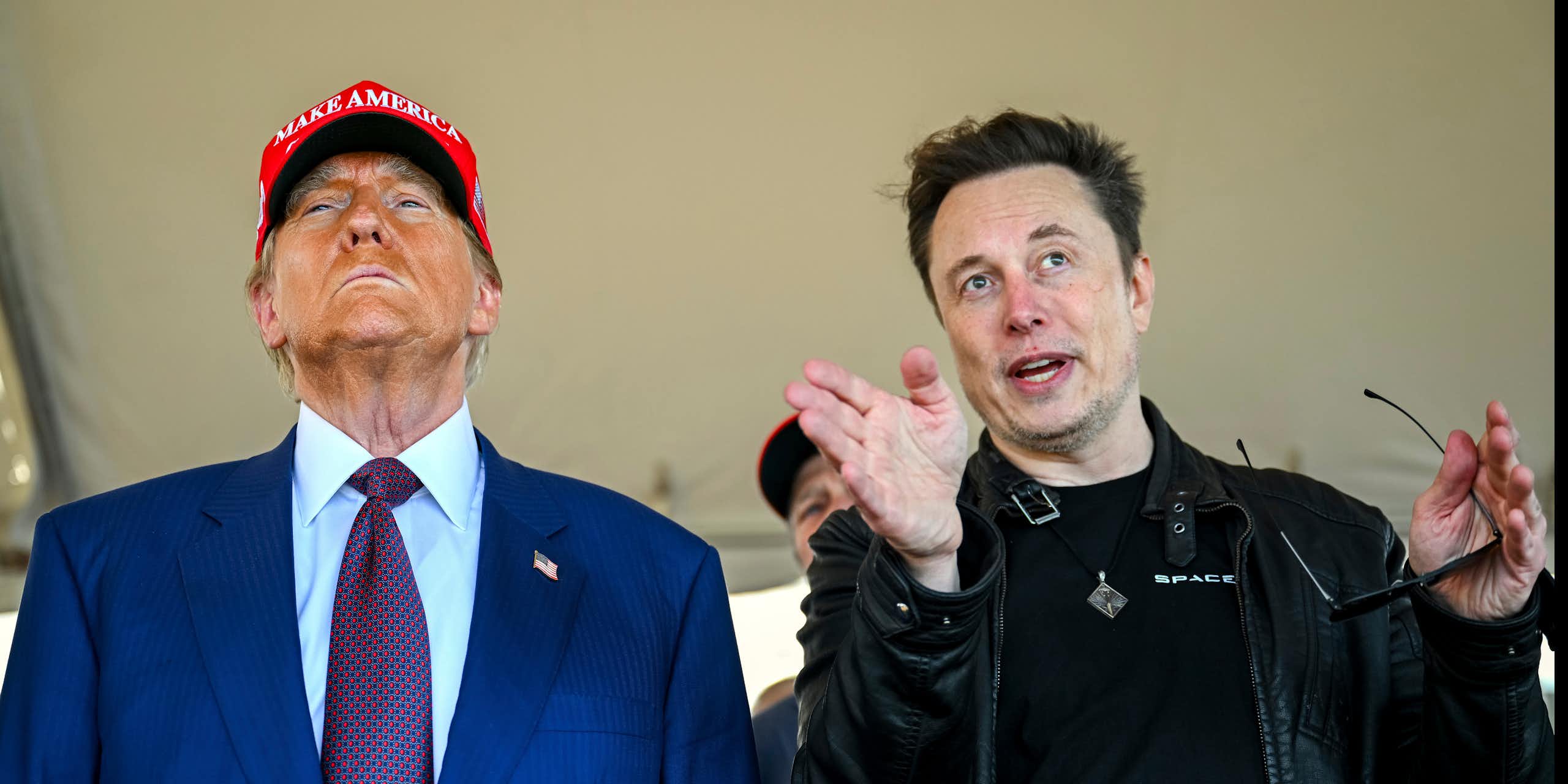 An older man with heavy facial bronzer wearing a red ball cap stands next to a younger man with dark hair who gestures as he looks ahead.
