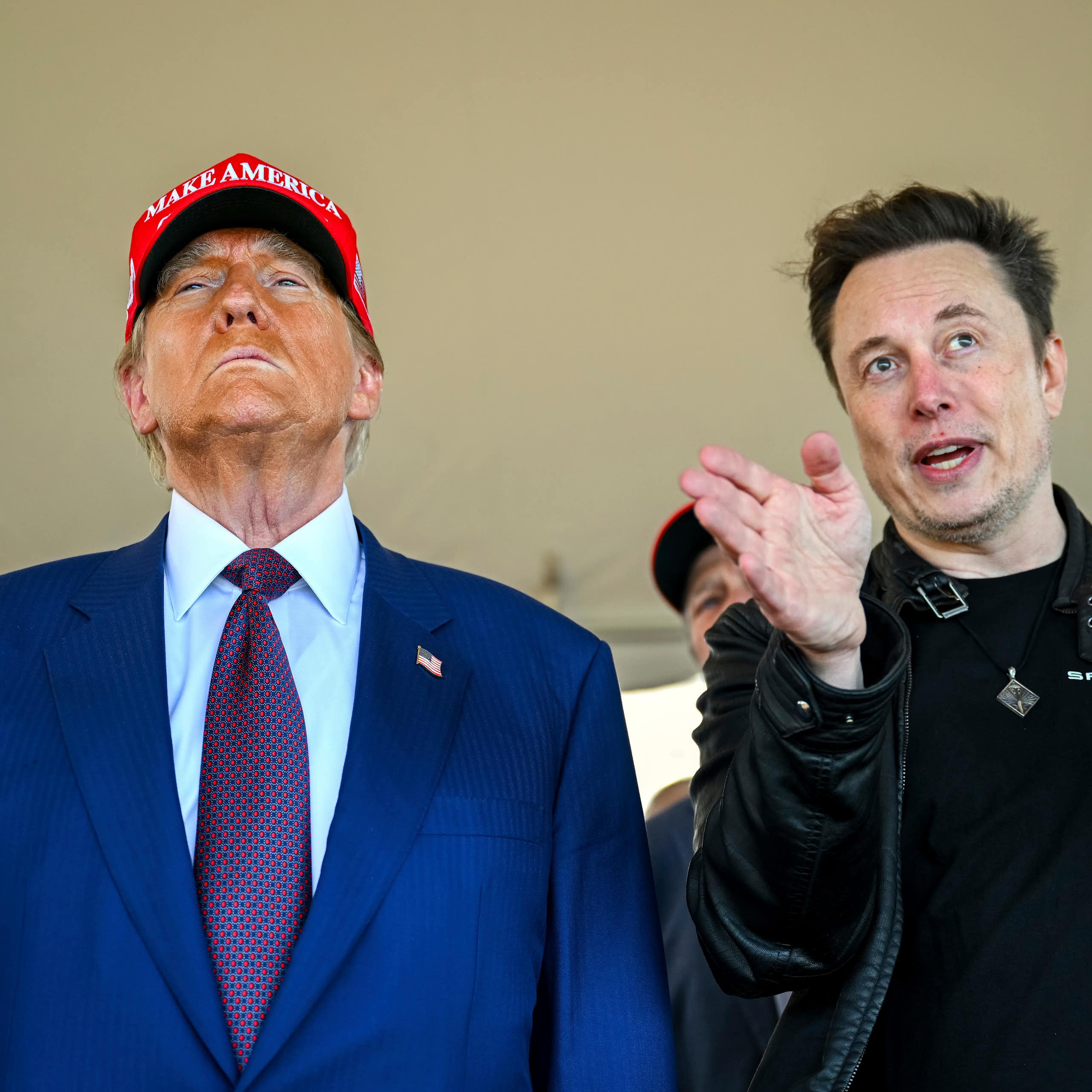 An older man with heavy facial bronzer wearing a red ball cap stands next to a younger man with dark hair who gestures as he looks ahead.