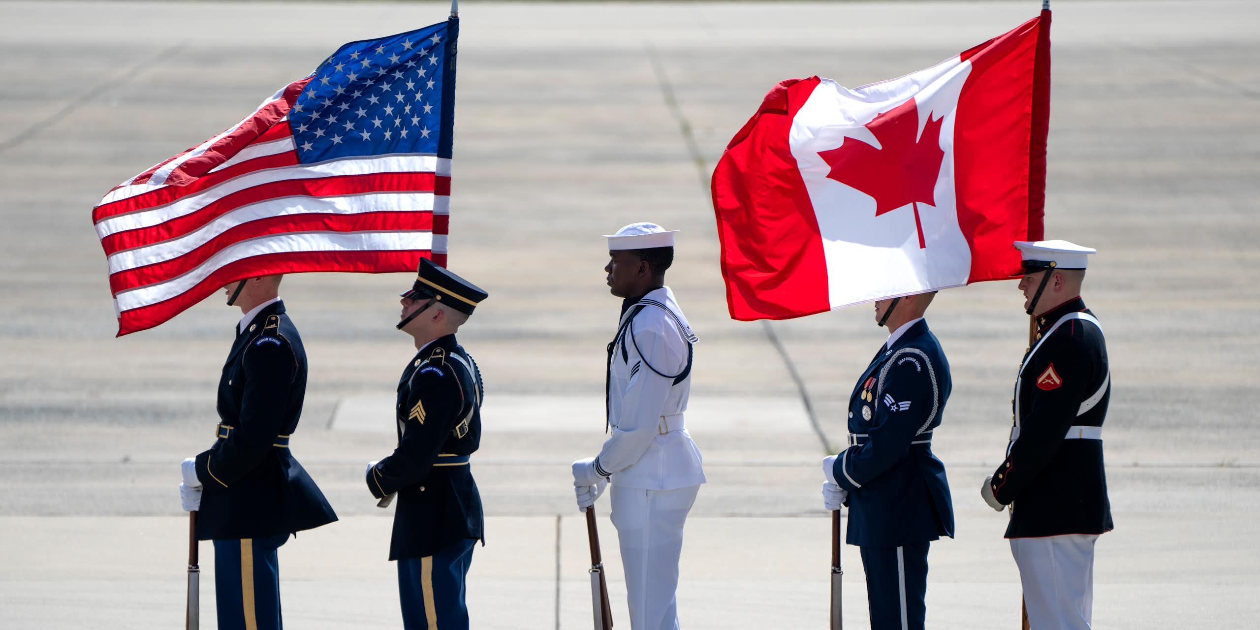 Honour guards stand on an airport tarmac holding an American and Canadian flag.