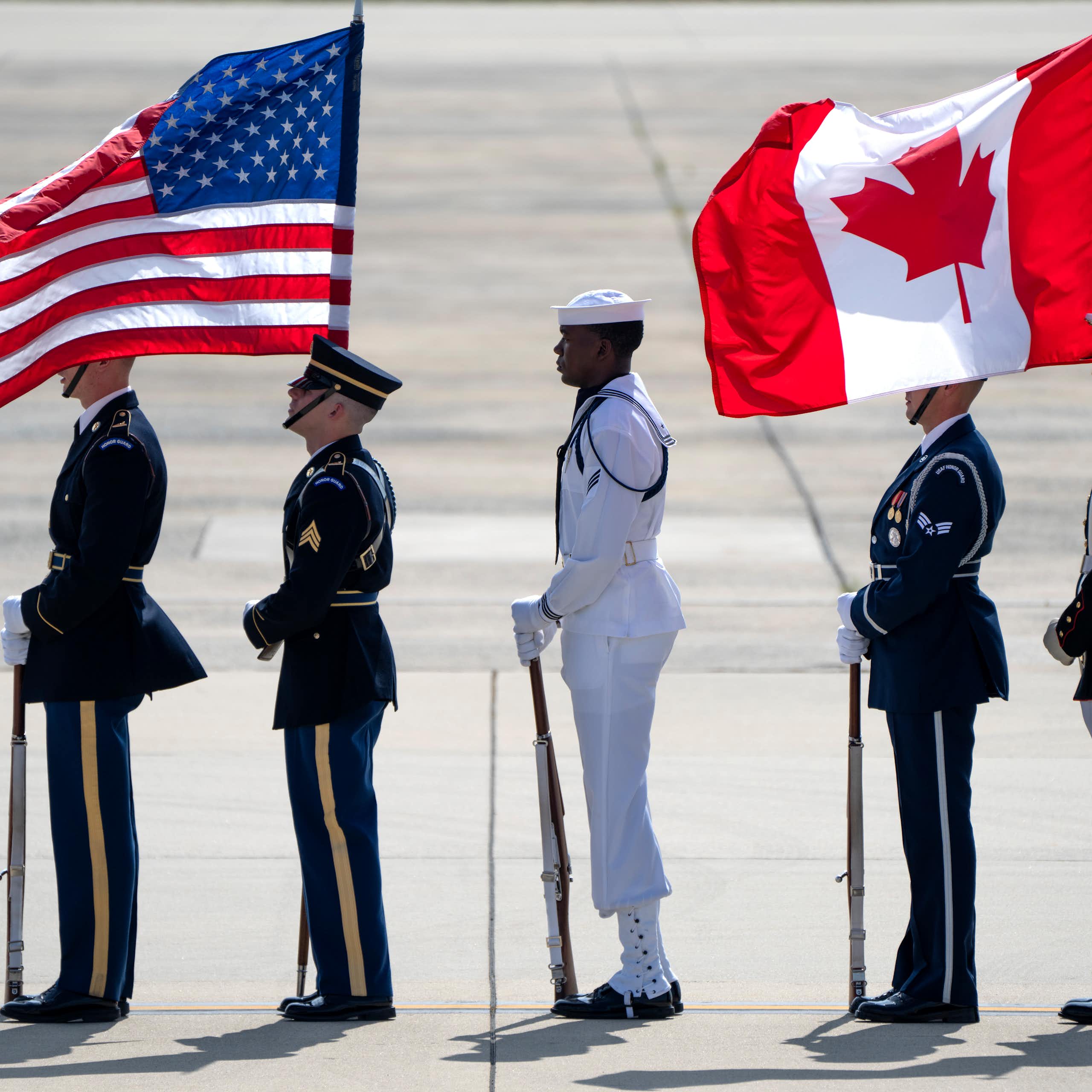 Honour guards stand on an airport tarmac holding an American and Canadian flag.