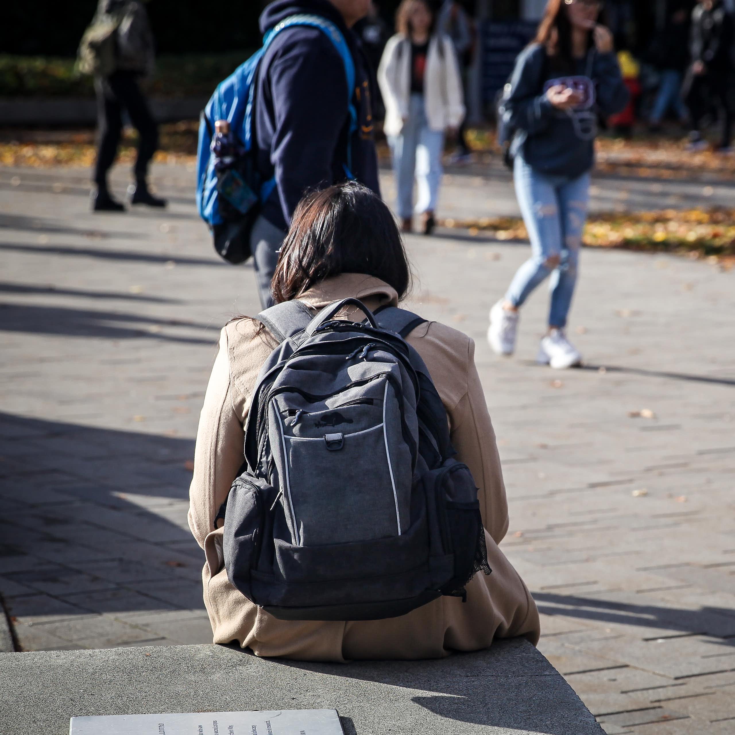 A woman with a backpack sits by a path as other people walk by