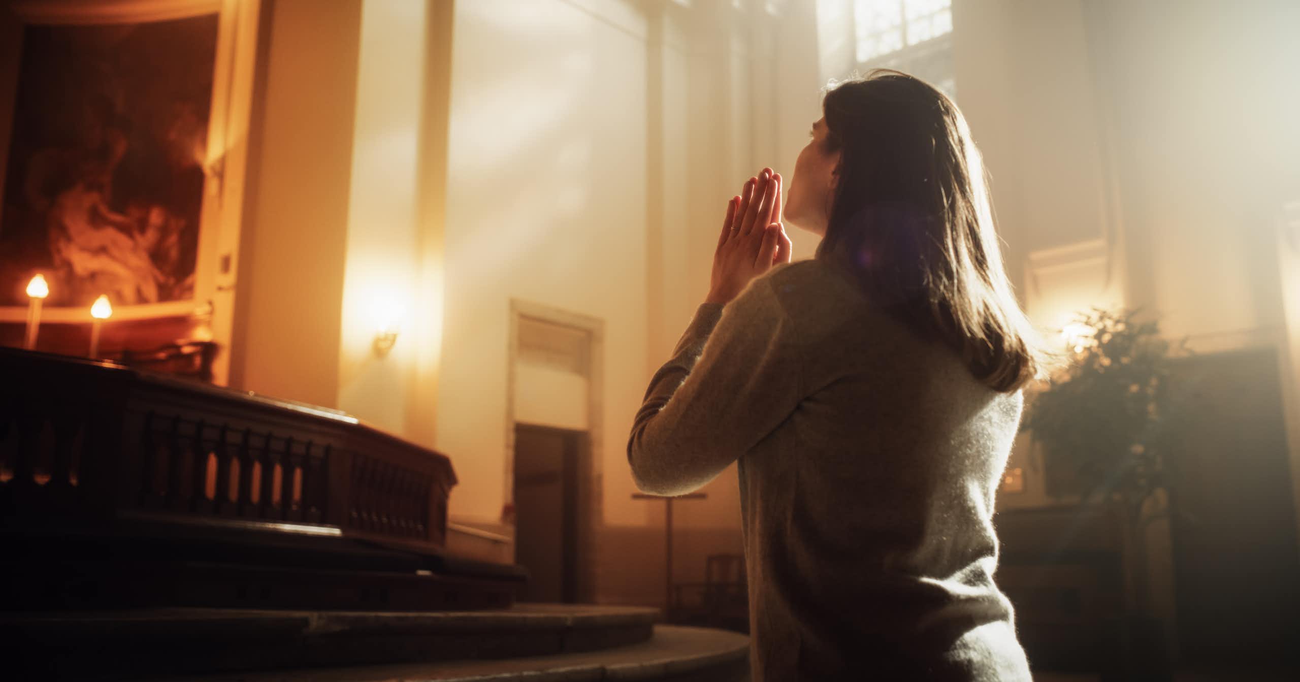 A woman in a church kneels and places her hands together in prayer.