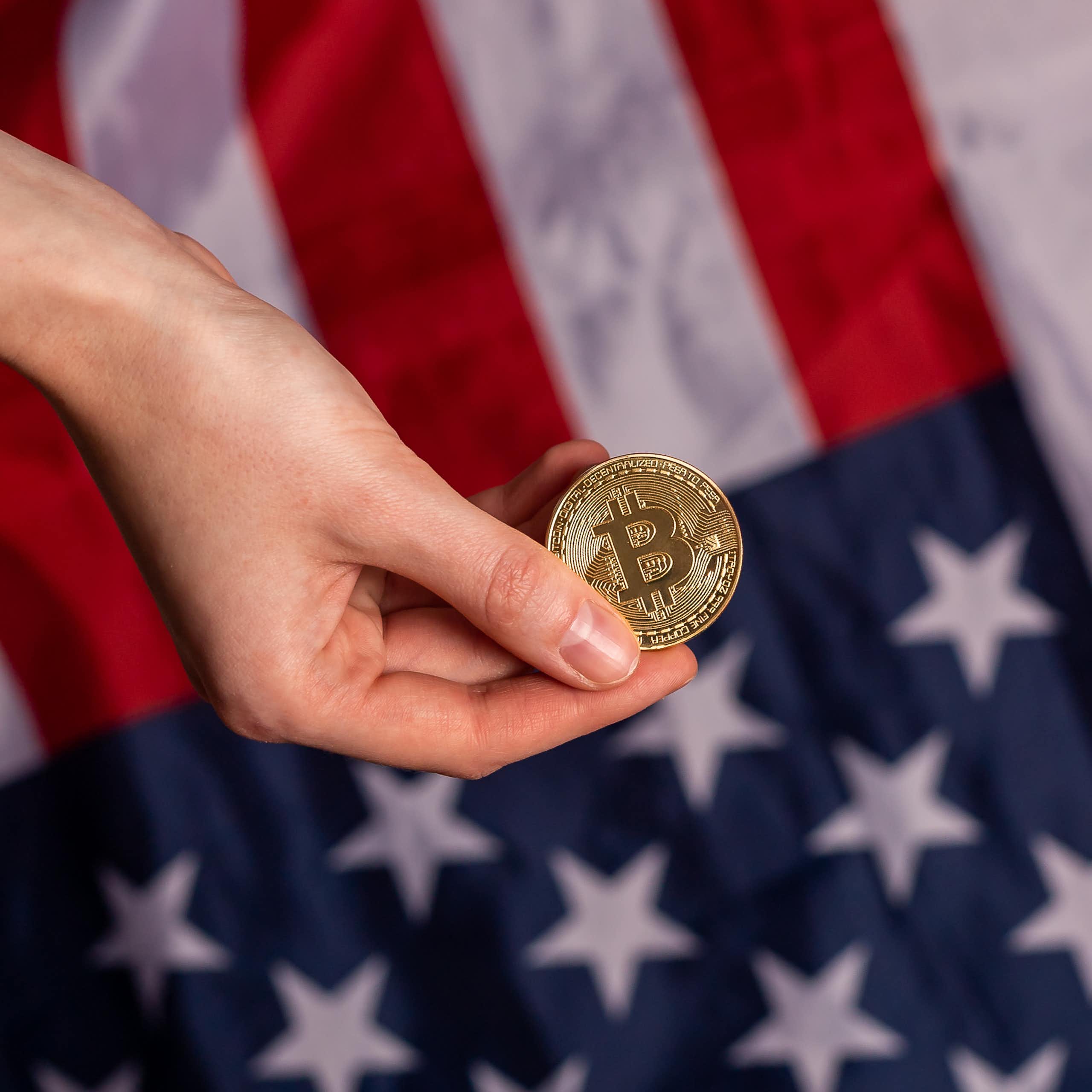 hand holding a representation of a bitcoin in front of the american flag