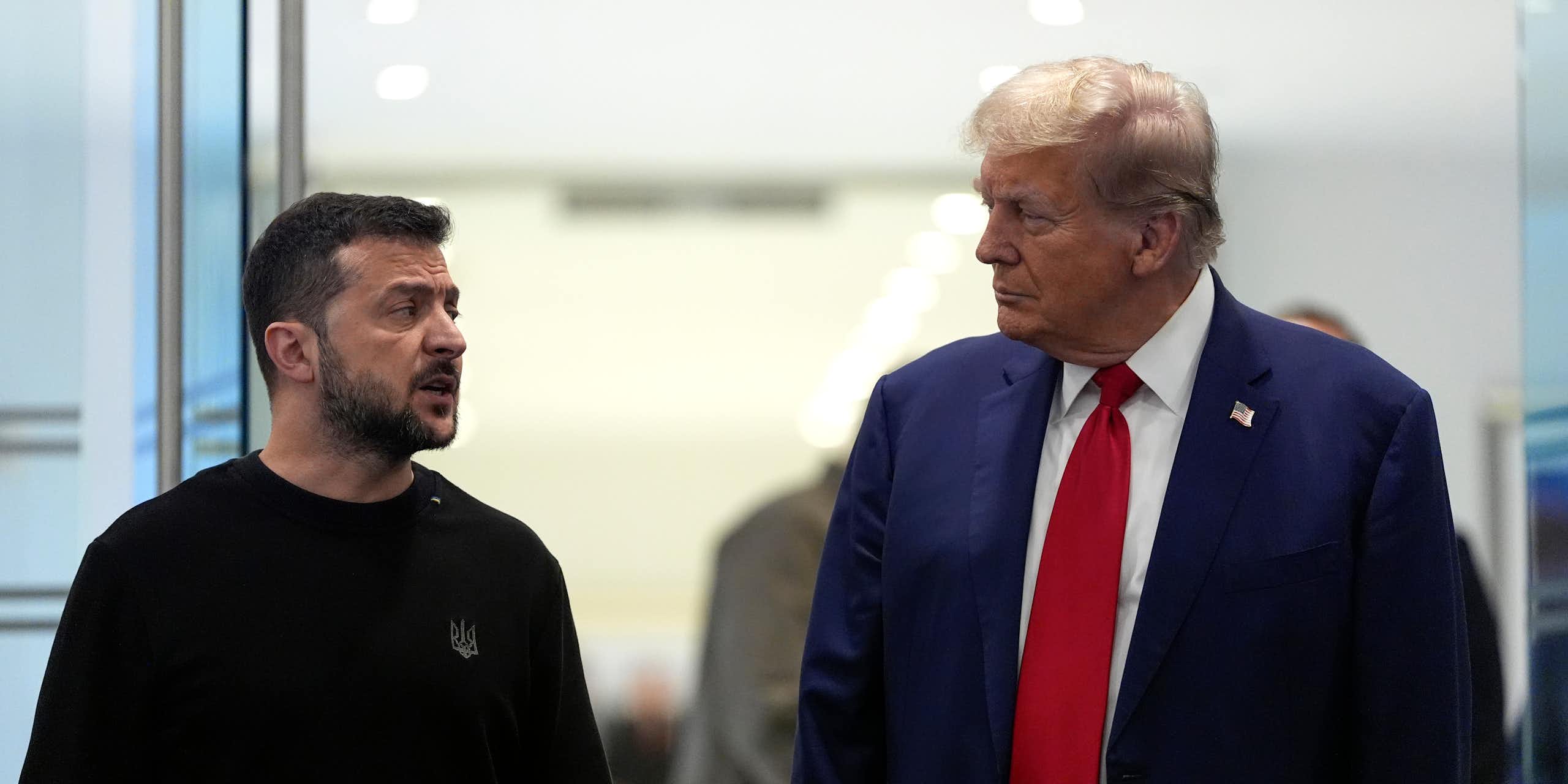 A rotund man in a blue suit and red tie with fluffy greyish blond hair talks to a shorter dark-haired man in a black shirt.