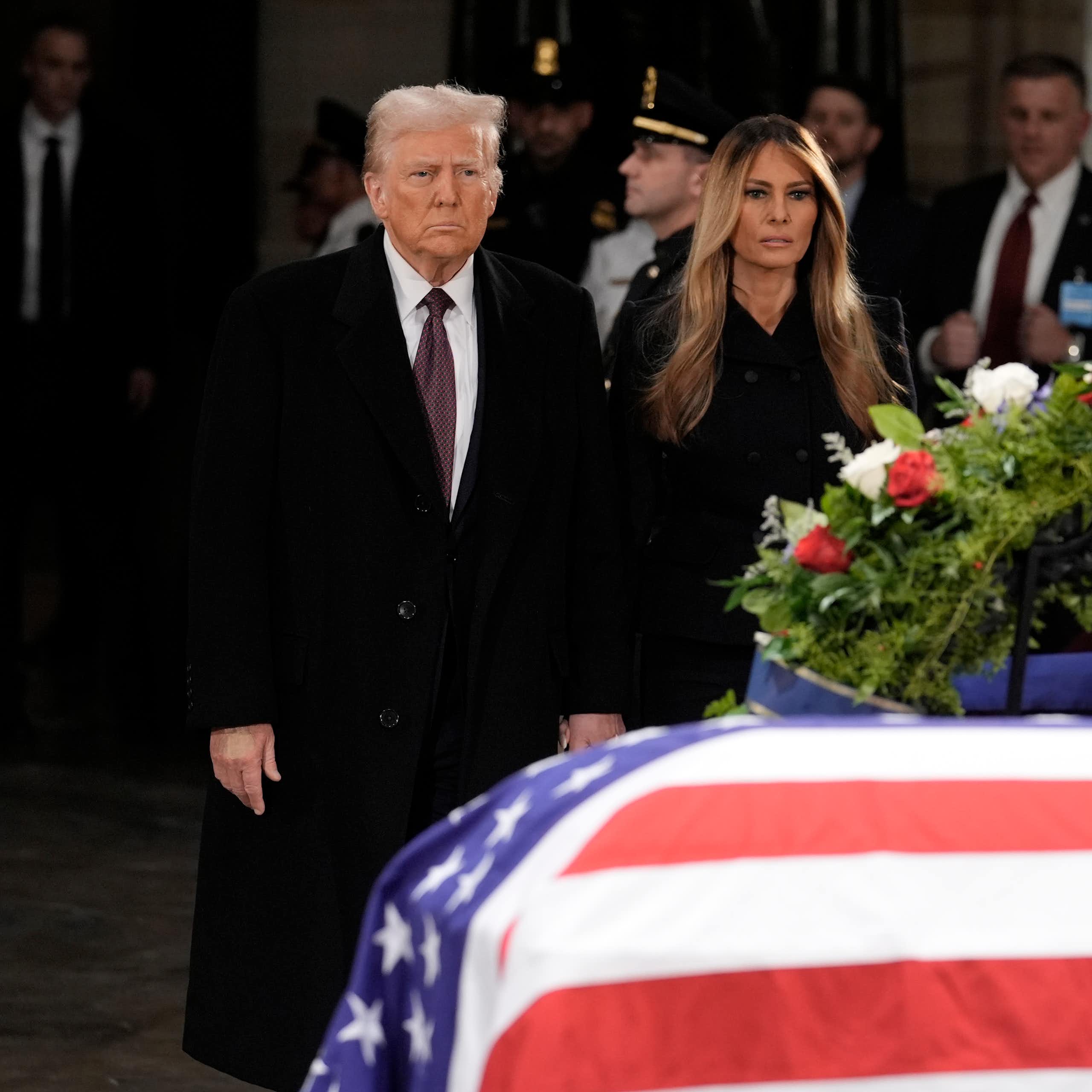A man with fluffy grey-blond hair stands next to a woman with long dark-blond hair behind a flag-draped casket.
