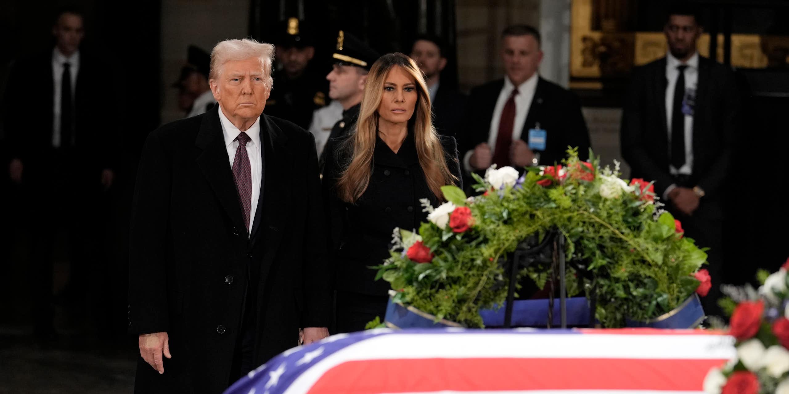 A man with fluffy grey-blond hair stands next to a woman with long dark-blond hair behind a flag-draped casket.
