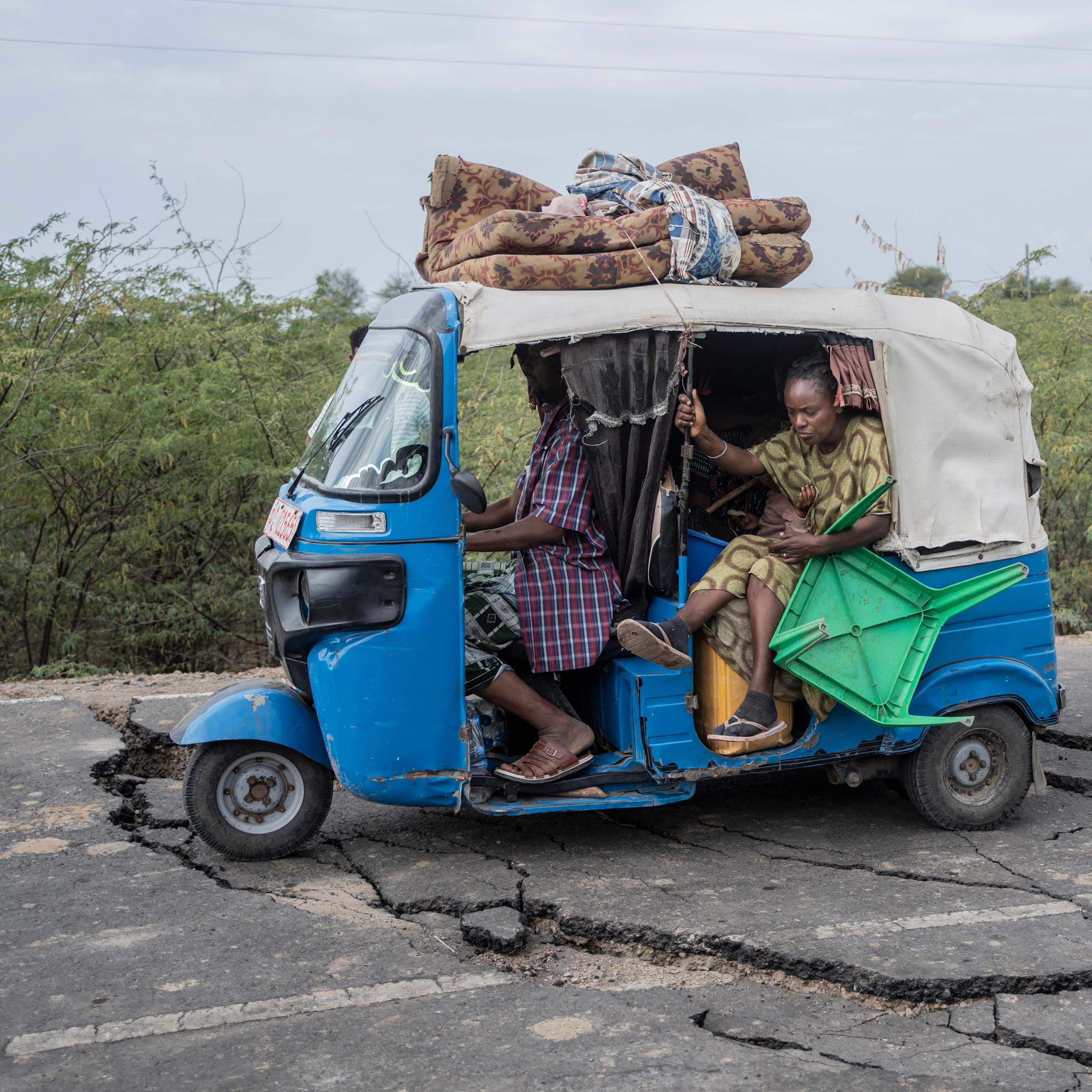A woman lifts her feet as the tiny blue and white vehicle she's traveling in navigates wide fissures in the road