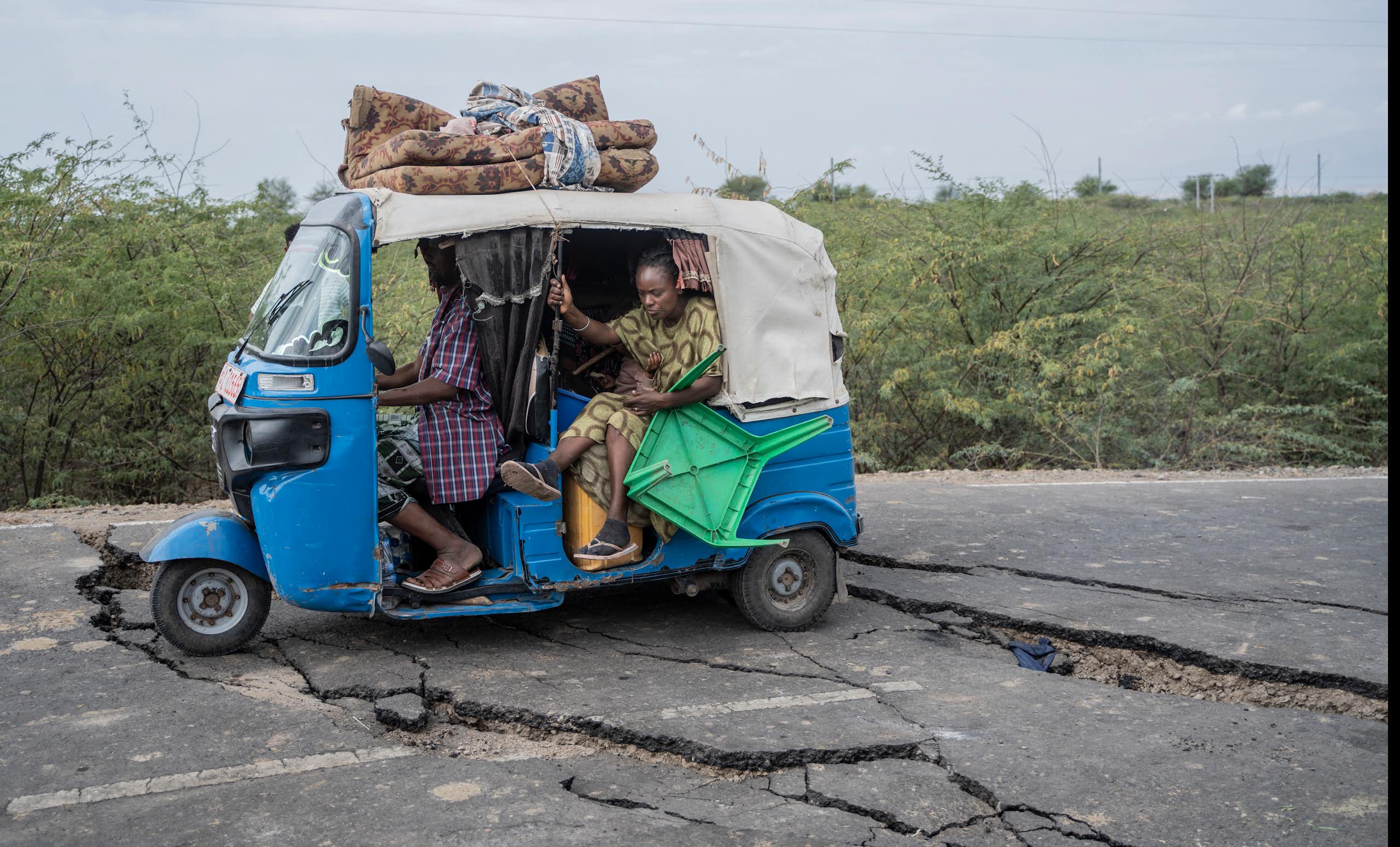 A woman lifts her feet as the tiny blue and white vehicle she's traveling in navigates wide fissures in the road