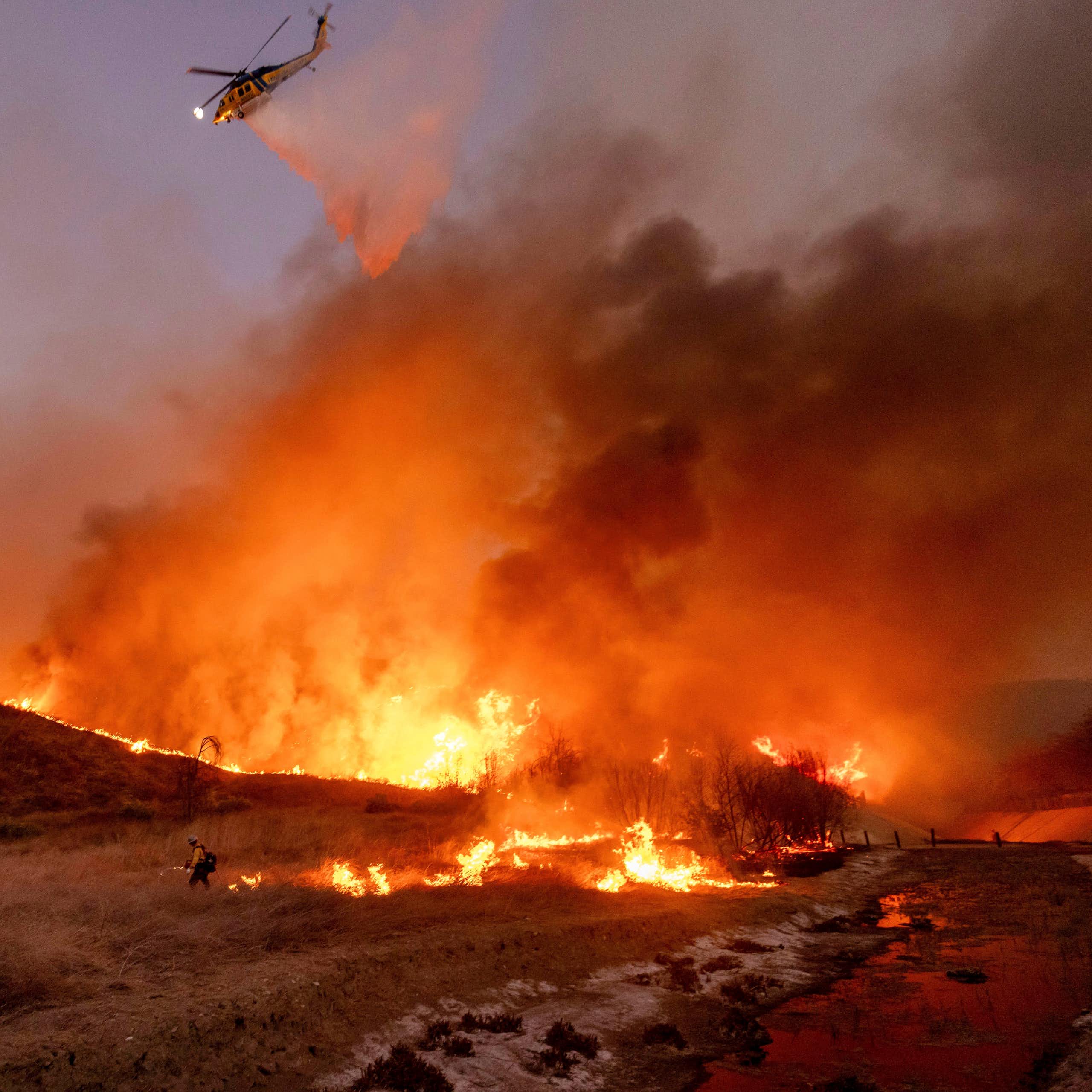 a large is burning on dry vegetation, two firefighters are on the ground and a helicopter flies above