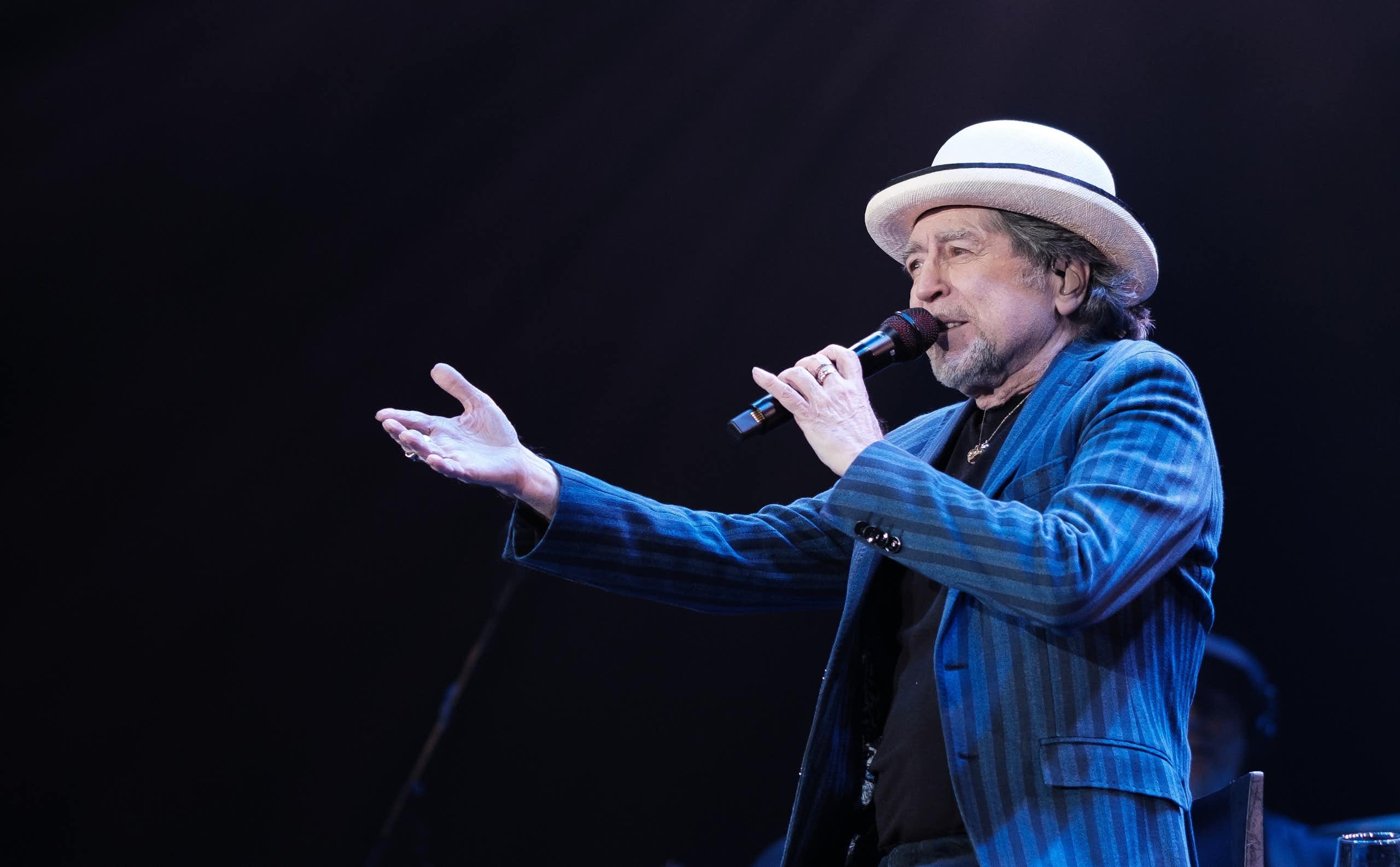 Un hombre con un sombrero y una americana azul canta sentado y señala al público.