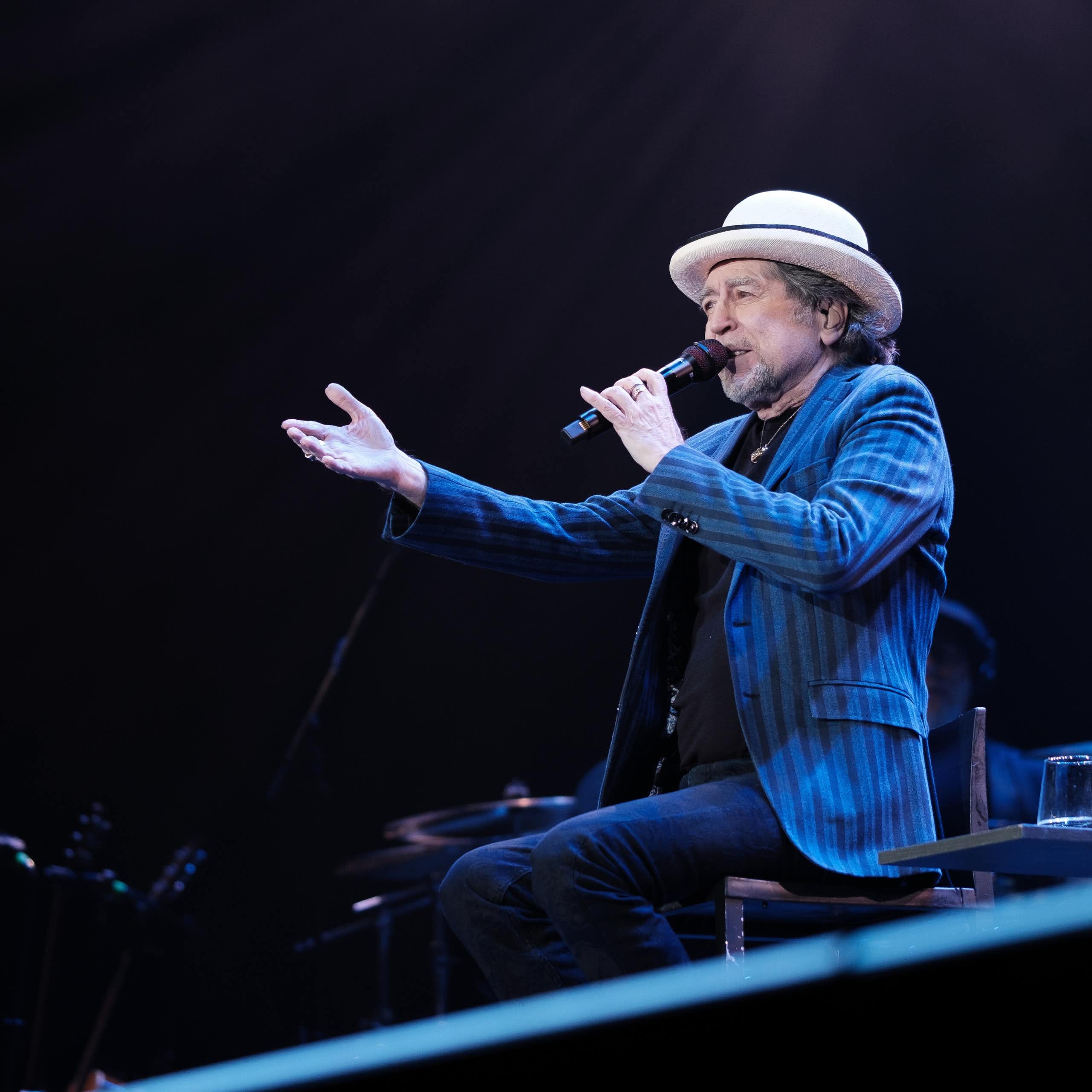Un hombre con un sombrero y una americana azul canta sentado y señala al público.