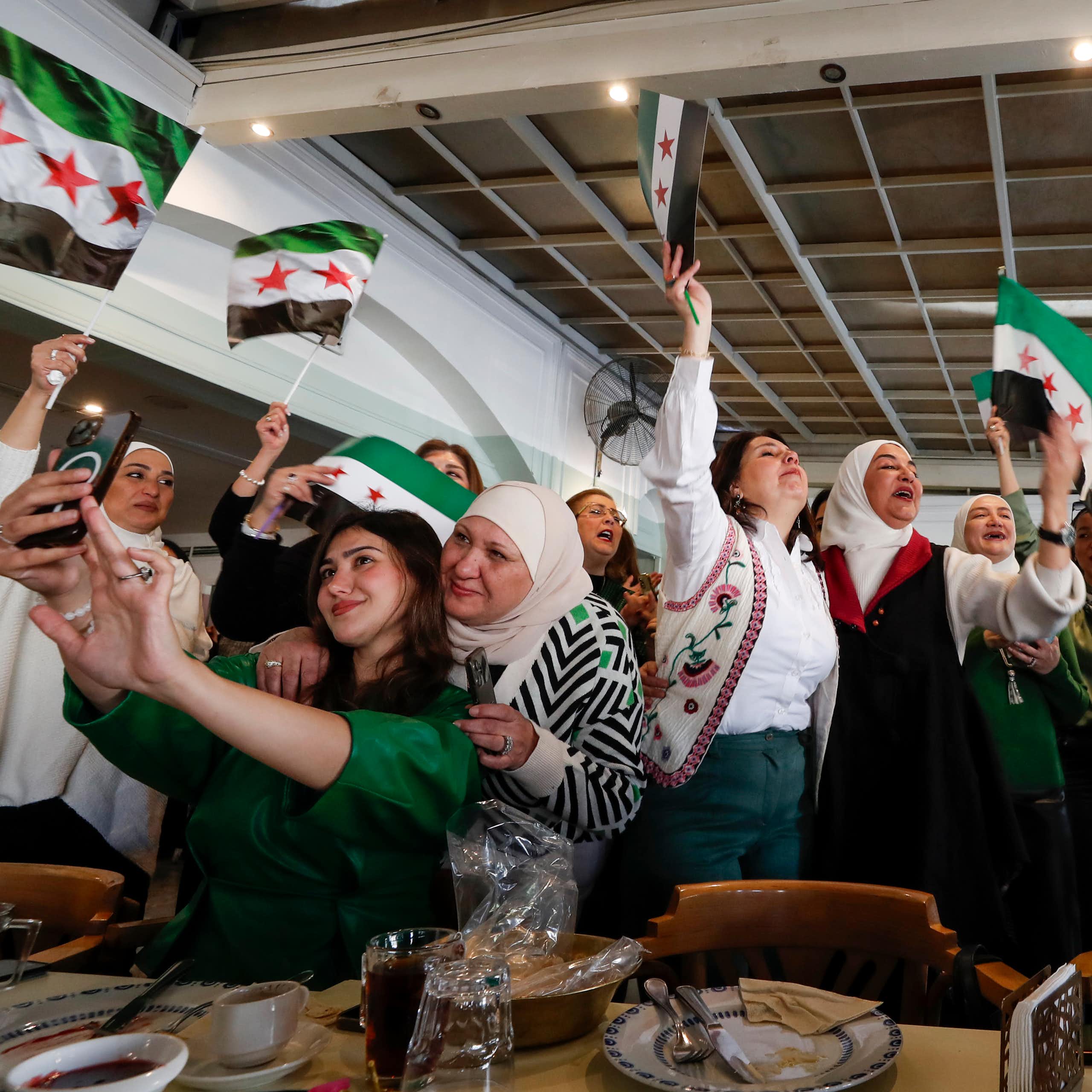 A group of women smile and celebrate, waving Syrian flags.