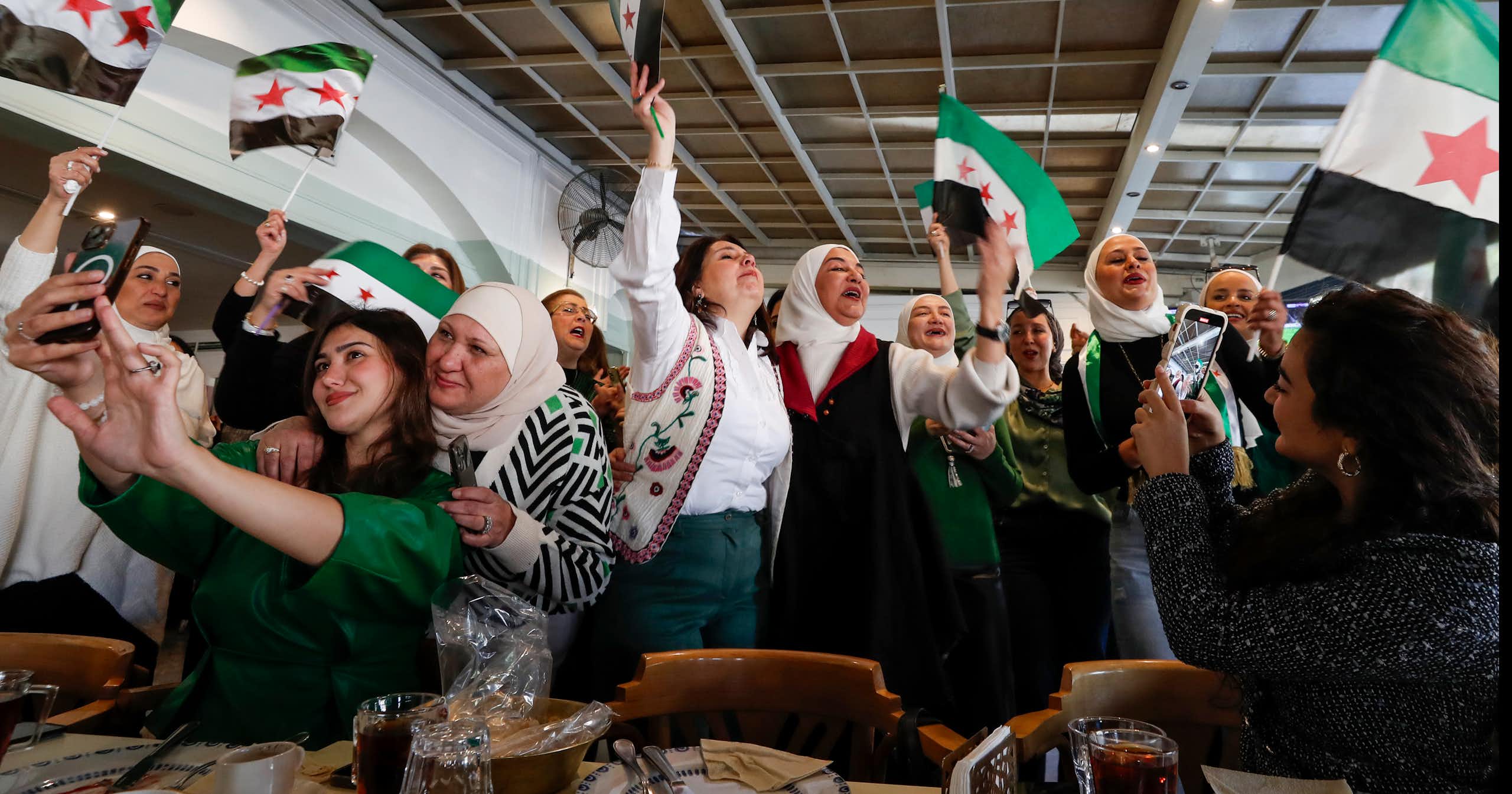 A group of women smile and celebrate, waving Syrian flags.