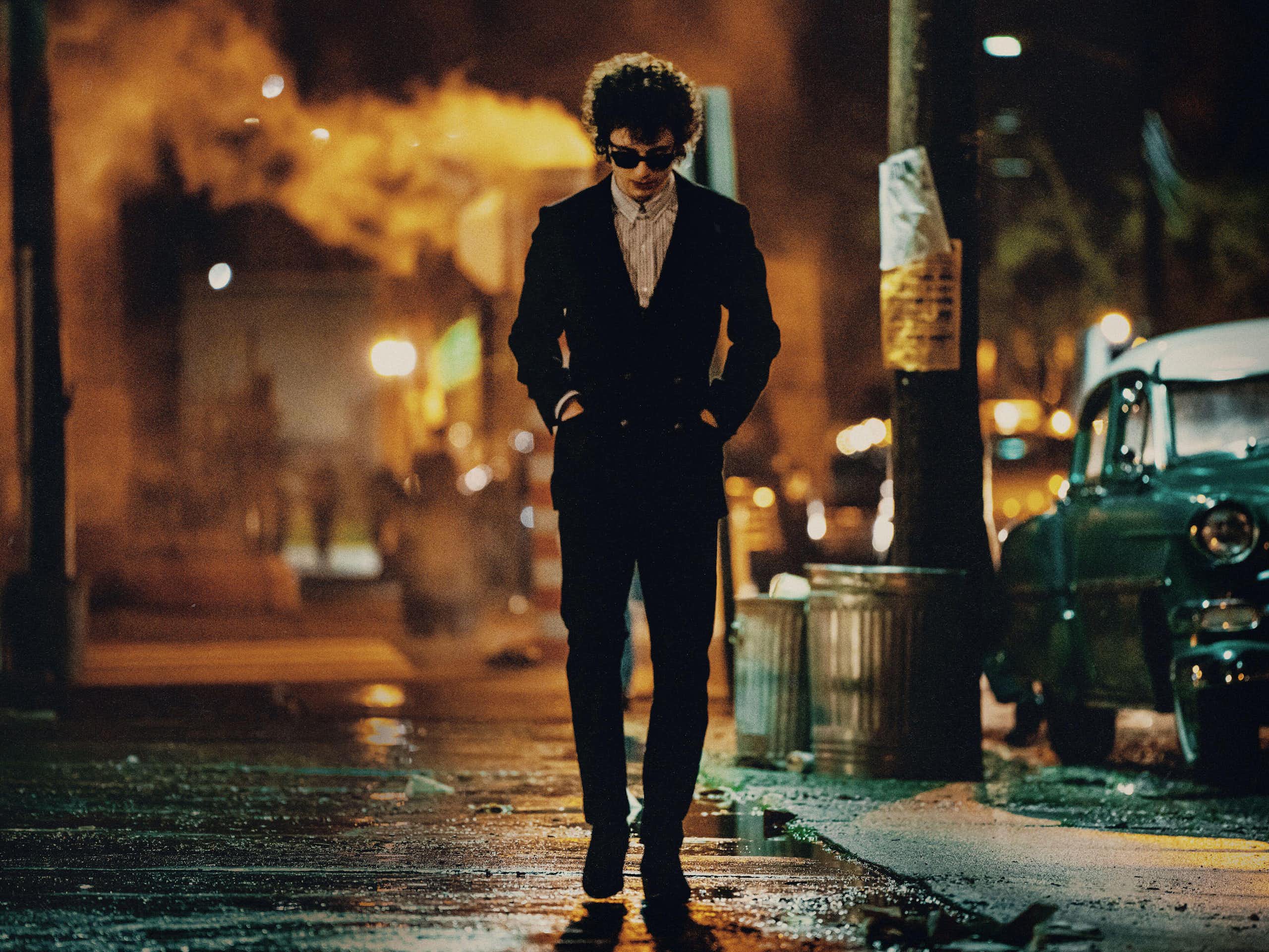 A lone male figure in sunglass and skinny suit walking down a 1960s NYC street at night