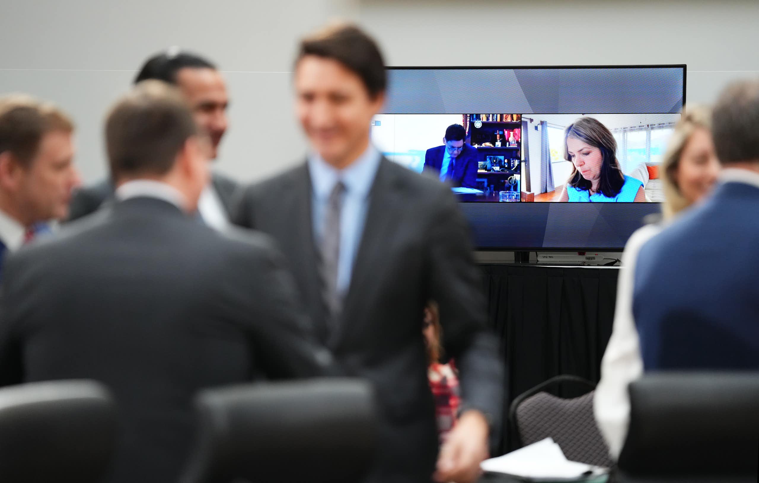 A woman in focus on a screen behind men in suits blurred standing in front of the screen.