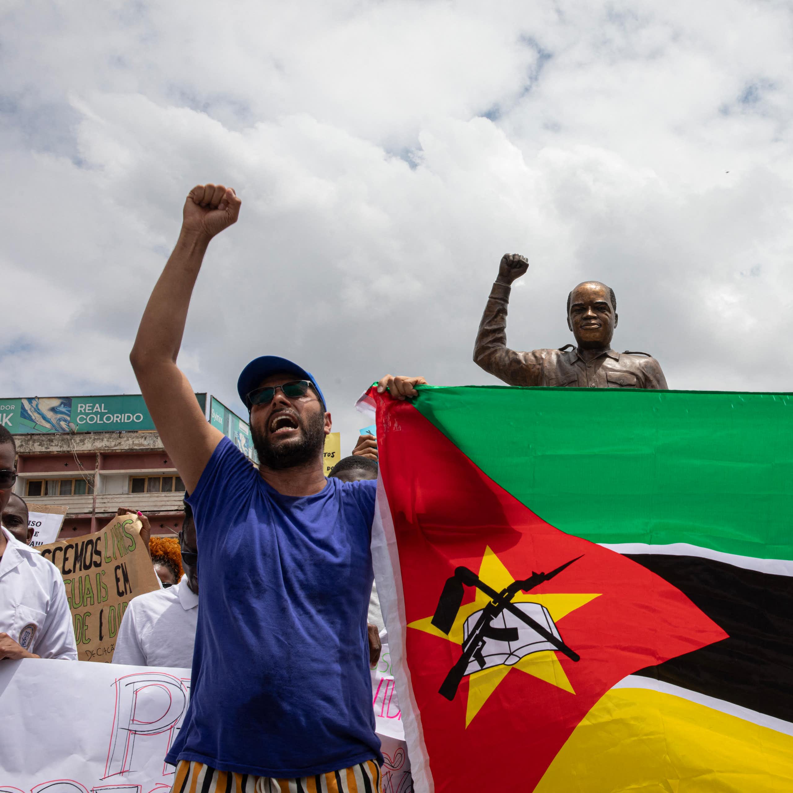 A man holding up a flag and raising his fist, in a gesture similar to a statue behind him