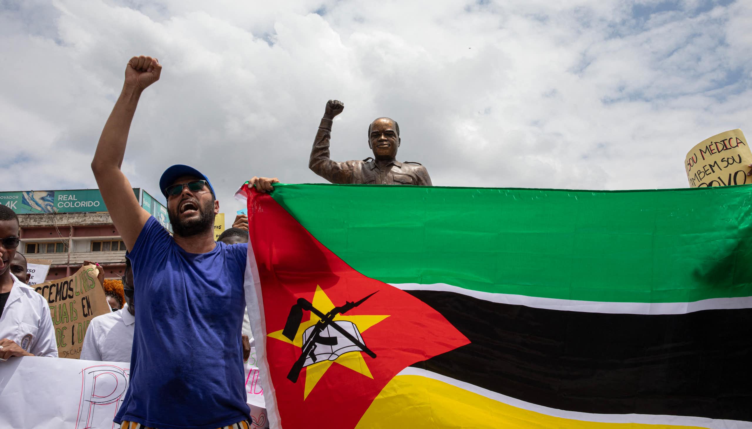 A man holding up a flag and raising his fist, in a gesture similar to a statue behind him