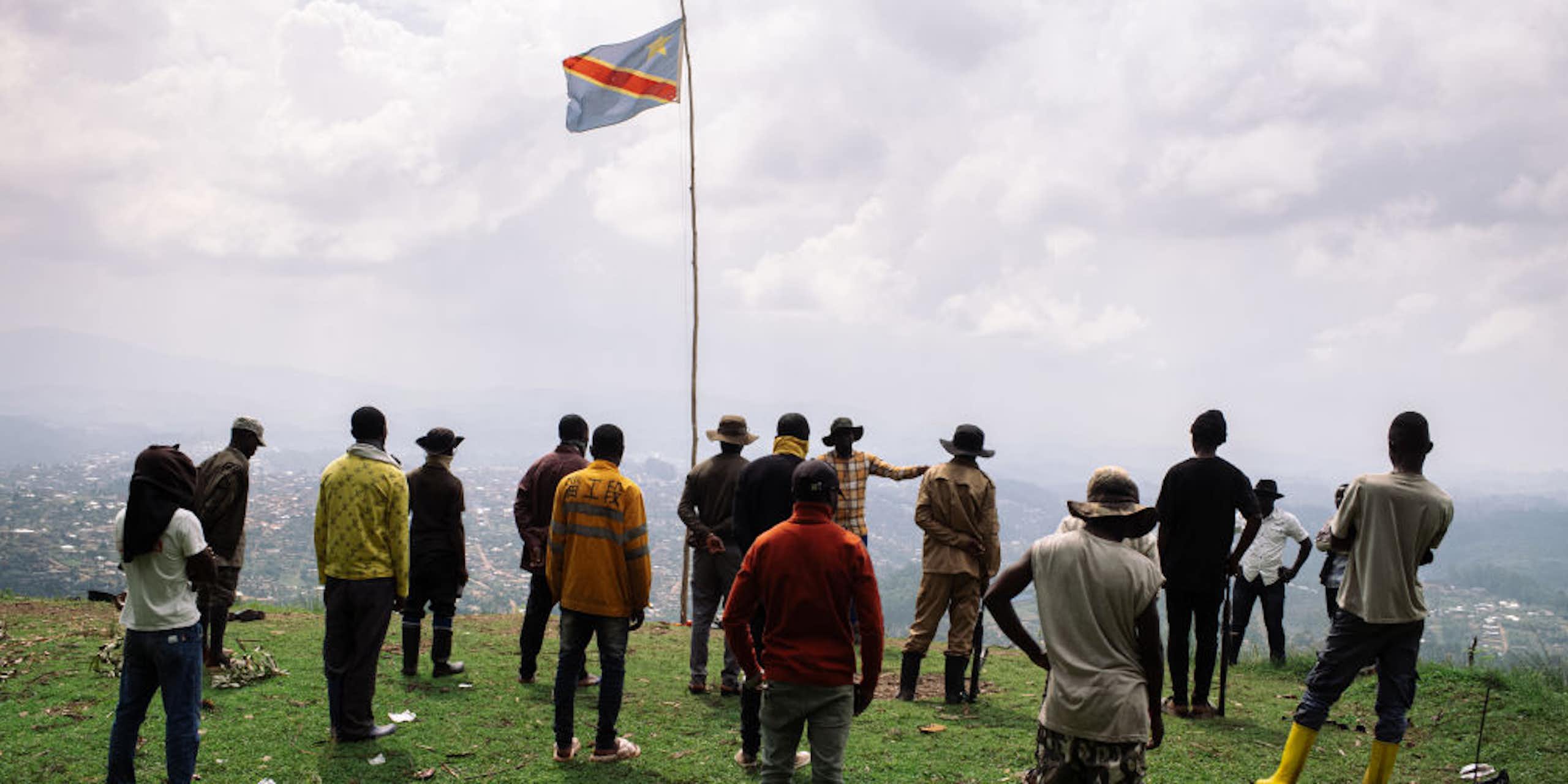 A group of standing men face a speaker standing on a hill overlooking a town below with the Congolese flag fluttering above them
