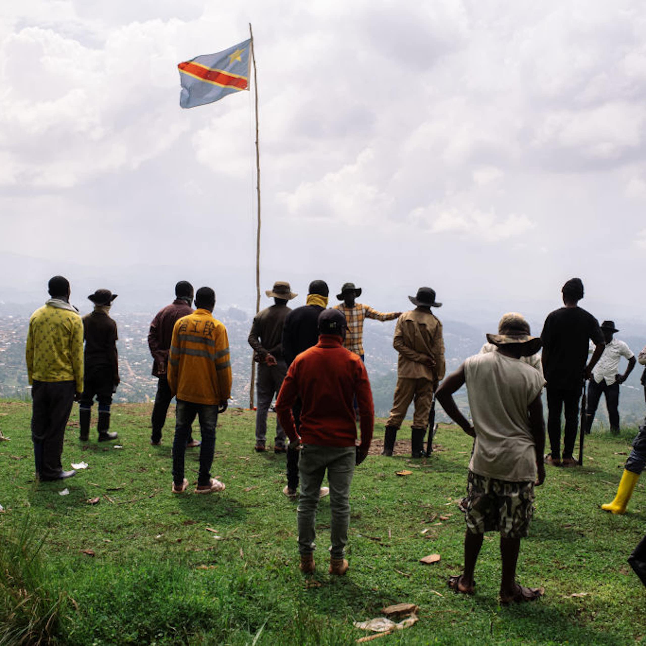 A group of standing men face a speaker standing on a hill overlooking a town below with the Congolese flag fluttering above them