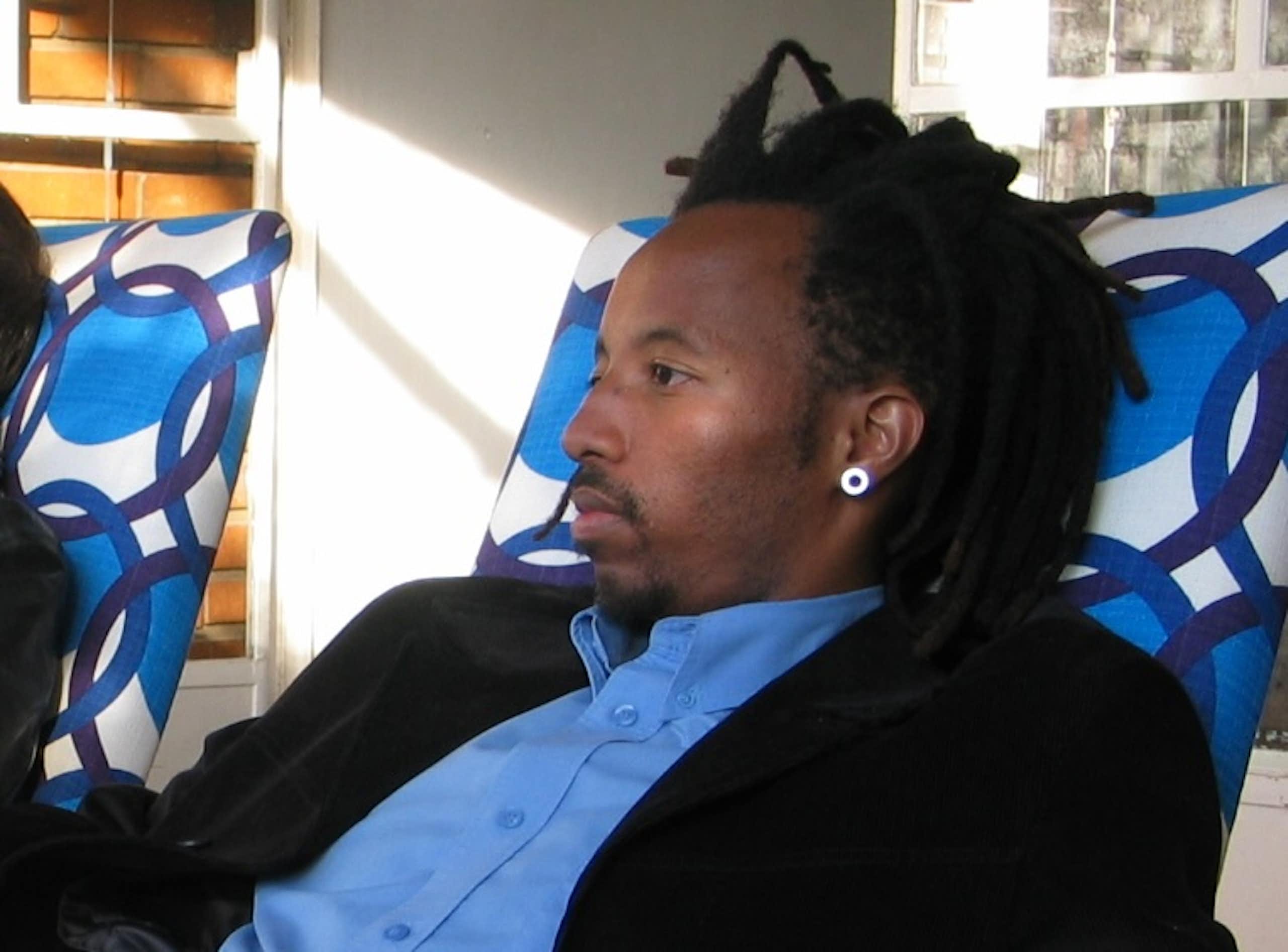 A young African man with dreadlocks reclines in a chair.
