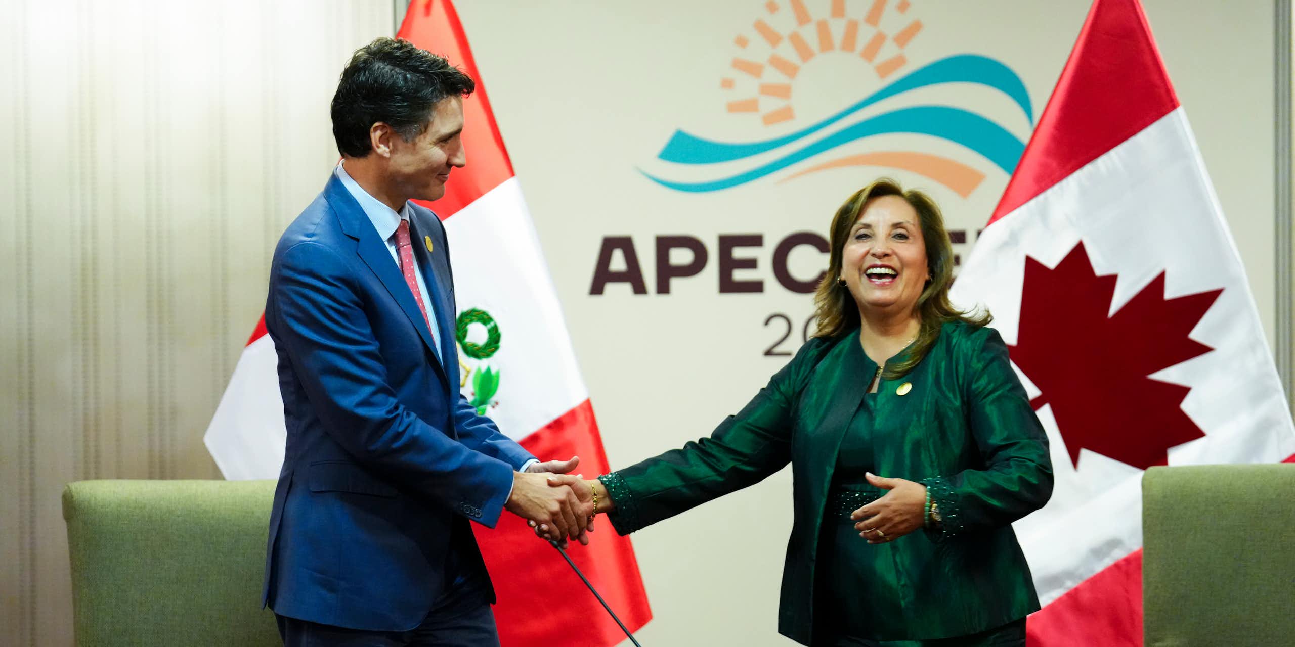 A dark-haired man in a navy suit shakes hands with a woman in a green suit.