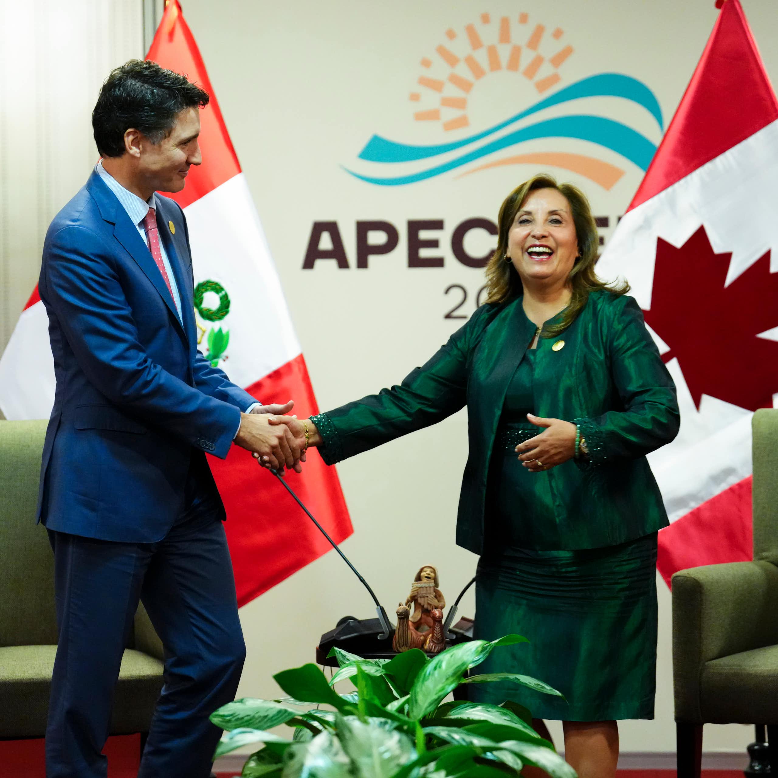 A dark-haired man in a navy suit shakes hands with a woman in a green suit.