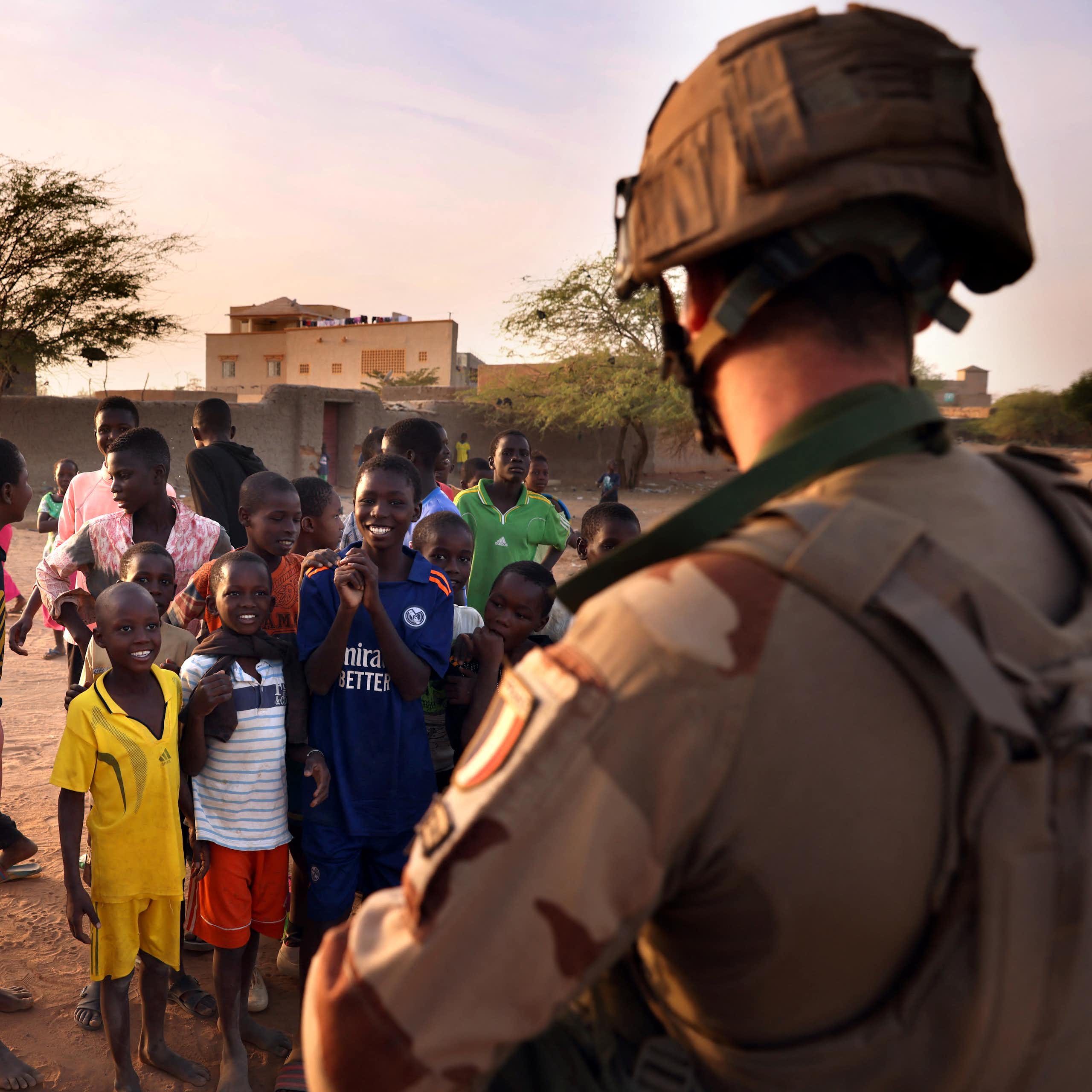 Children play soccer and stand looking at a man in military uniform