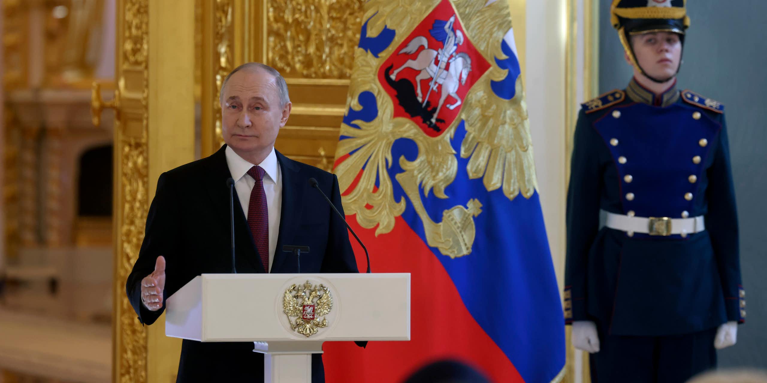 Vladimir Putin stands at a lectern flanked by a ceremonial guardsman.