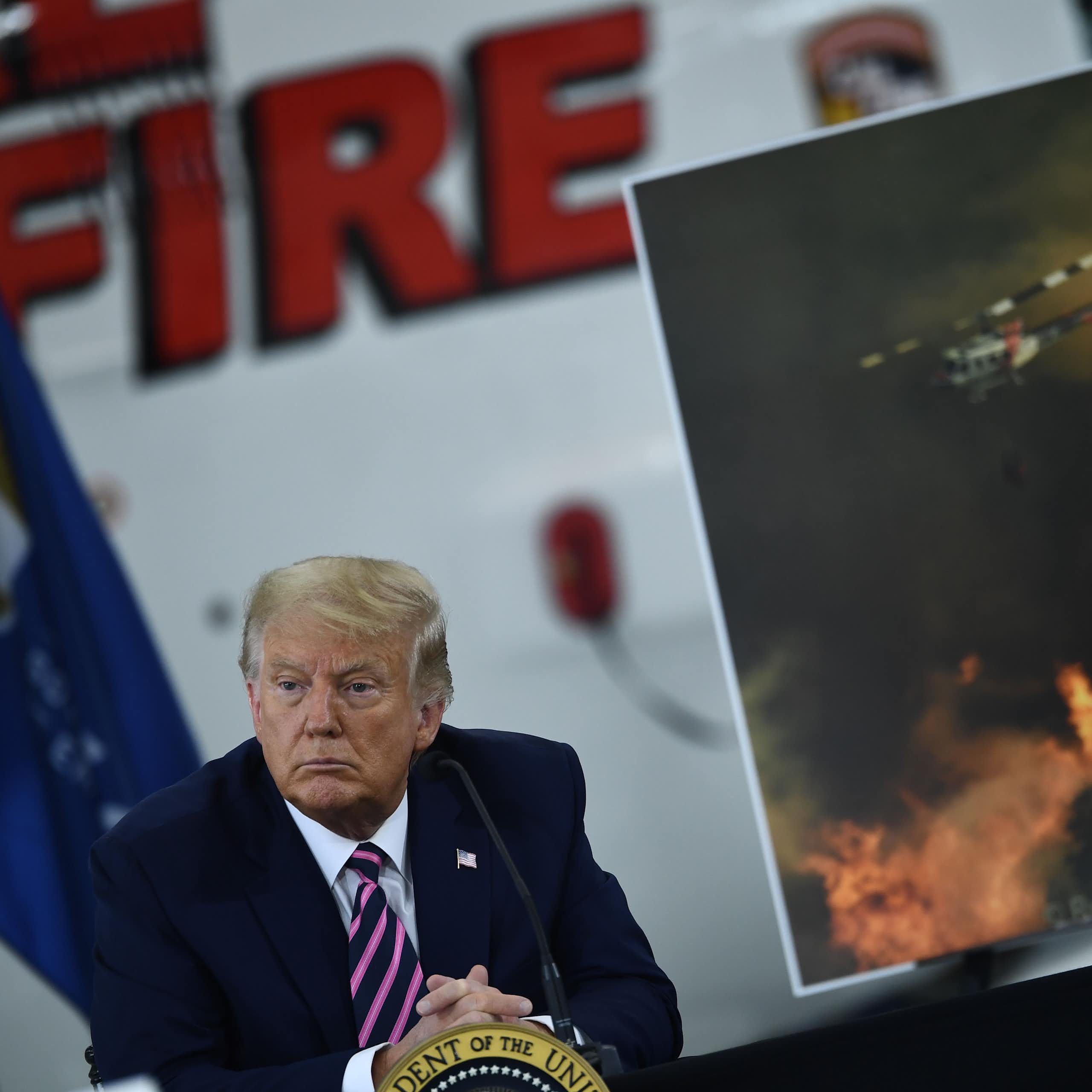 Trump está sentado em uma reunião sobre incêndios florestais, com uma imagem de um helicóptero jogando água em um incêndio ao fundo.