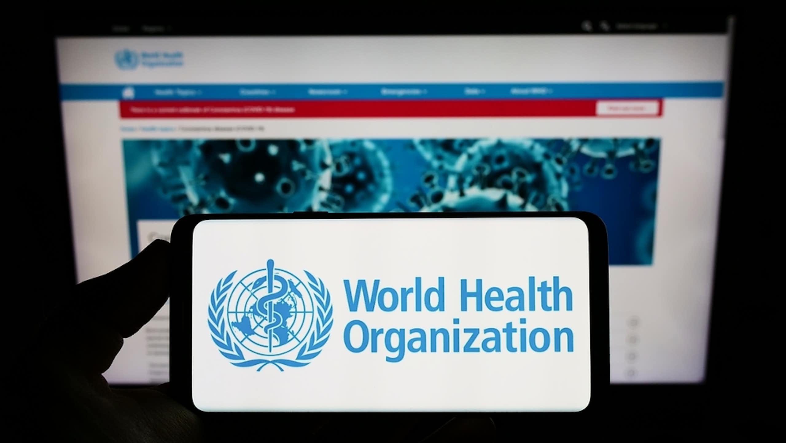 Smartphone with WHO logo held against computer screen showing WHO website
